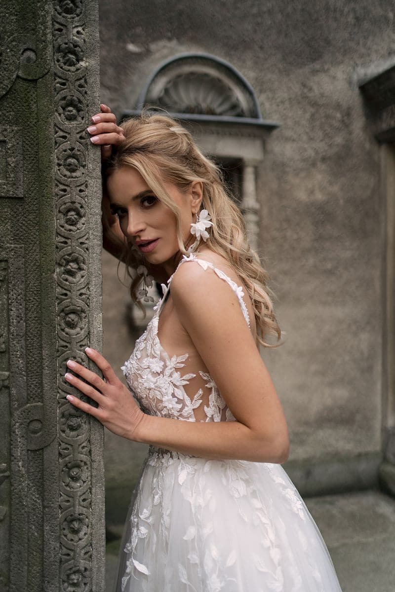 Eine Frau im Dama Couture Samira Hochzeitskleid mit weißer, geblümter Spitze posiert an einer Steinsäule mit langem, gewelltem blondem Haar und blickt in die Kamera. Hinter ihr befinden sich eine verzierte, verwitterte Steinmauer und ein gewölbtes Fenster.