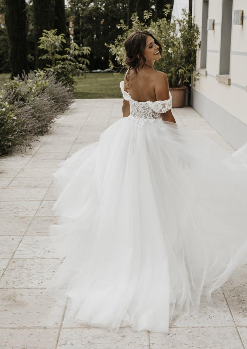 Eine Frau in einem Bridentity Enchantment Hochzeitskleid steht auf einem steinernen Gehweg im Freien, umgeben von Grün, schaut zurück und lächelt in ihrem schulterfreien weißen Brautkleid.