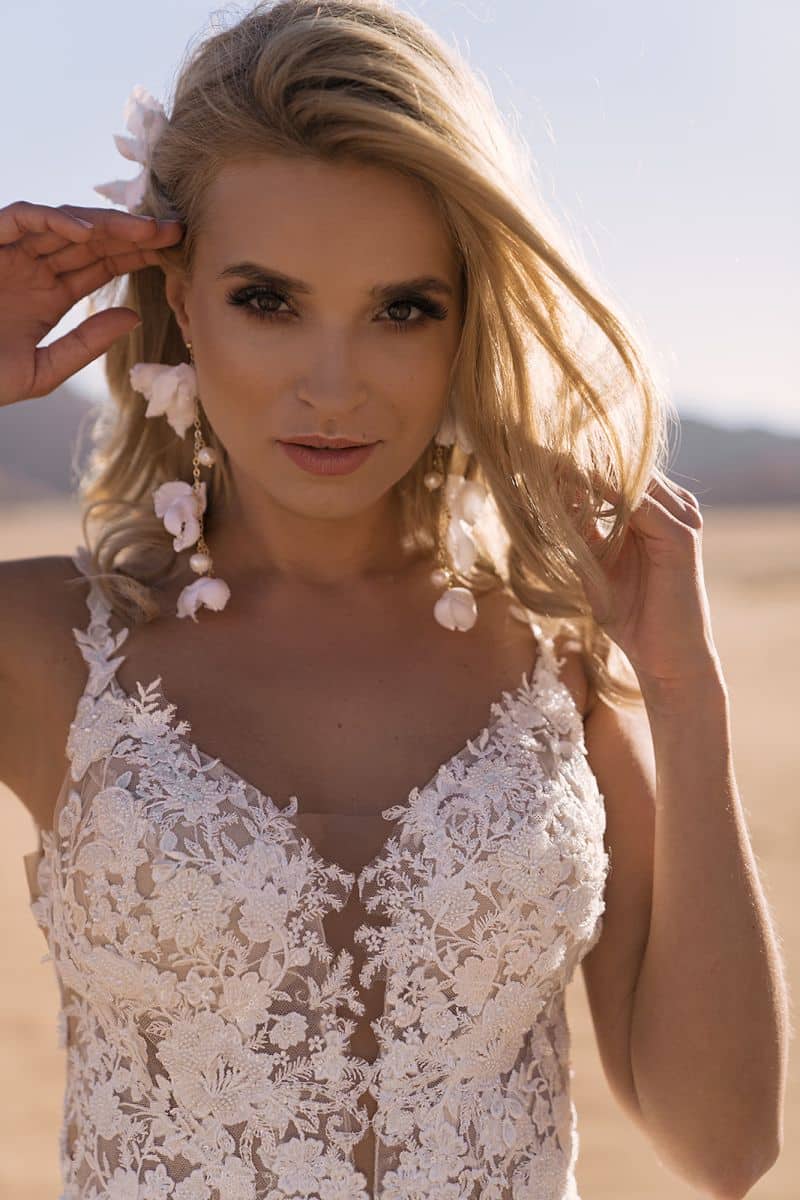 Eine Frau im Dama Couture Gilly Brautkleid steht im Freien im Sonnenlicht und schaut in die Kamera. Sie berührt ihr langes blondes Haar und ihre weißen Blumenohrringe; der sandige Fleck hinter ihr unterstreicht diesen stilvollen Brautmoden-Moment.