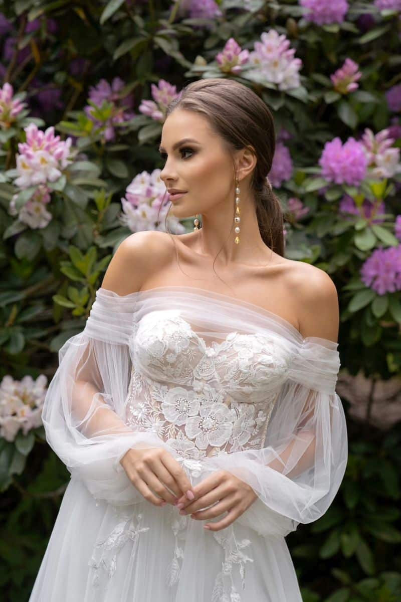 Eine Frau im Dama Couture Lanka Hochzeitskleid - ein schulterfreies, spitzenbesticktes Hochzeitskleid - steht vor blühenden rosa und weißen Blumen, ihr Haar ist nach hinten gestylt und lange Ohrringe vervollständigen ihren Brautlook.