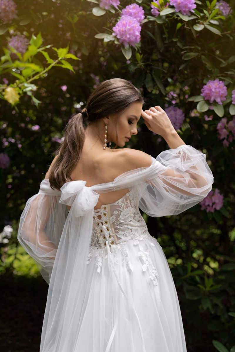 Eine Frau trägt das Dama Couture Lanka Hochzeitskleid und steht im Freien zwischen grünem Laub und lila Blüten, dreht ihren Kopf zur Seite und legt sanft die Hand an ihr Gesicht.