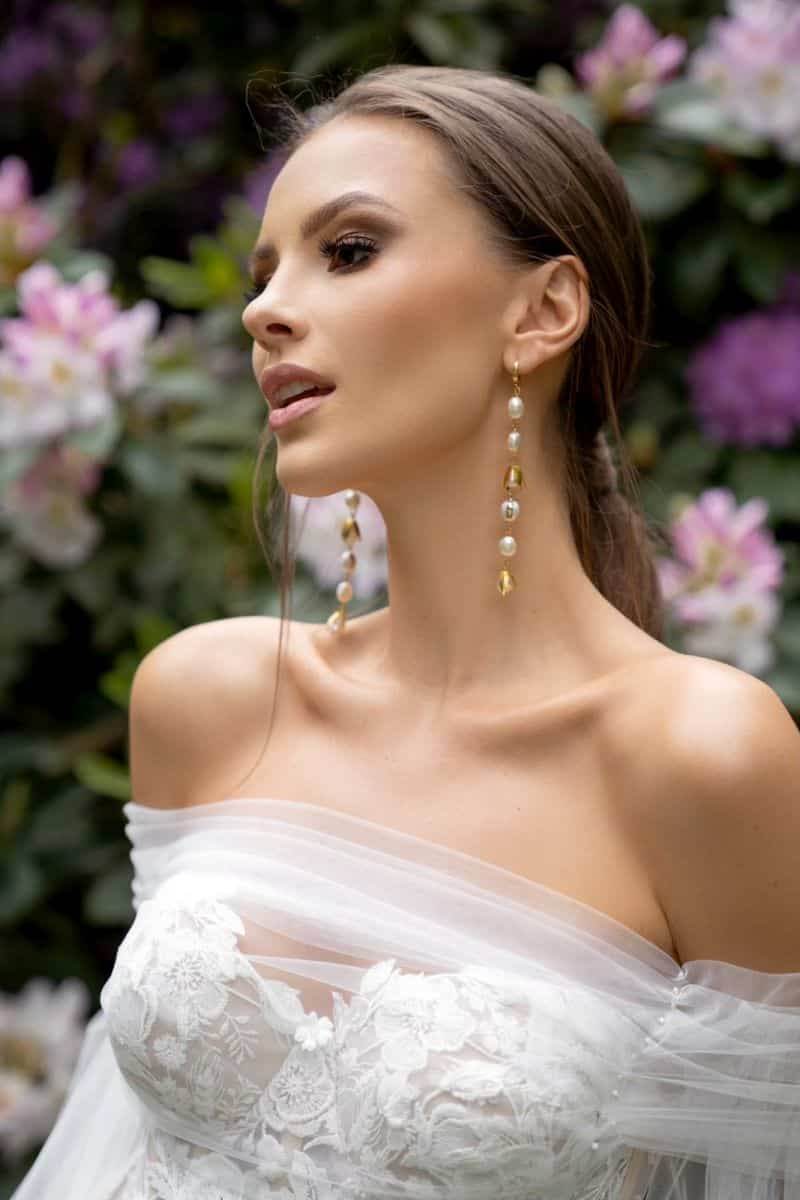 Eine Frau, die das Dama Couture Lanka Hochzeitskleid mit Spitzendetails und langen Ohrringen trägt, posiert im Freien zwischen blühenden rosa und weißen Blumen.