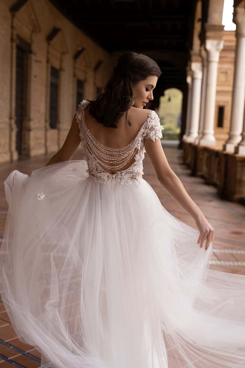 Eine Frau im Dama Couture Pavetta Brautkleid mit fließender weißer Silhouette, tiefem Rücken und Perlenketten steht auf einem überdachten Spaziergang im Freien, blickt über ihre Schulter und hält ihren Rock fest - moderne Braut-Eleganz.