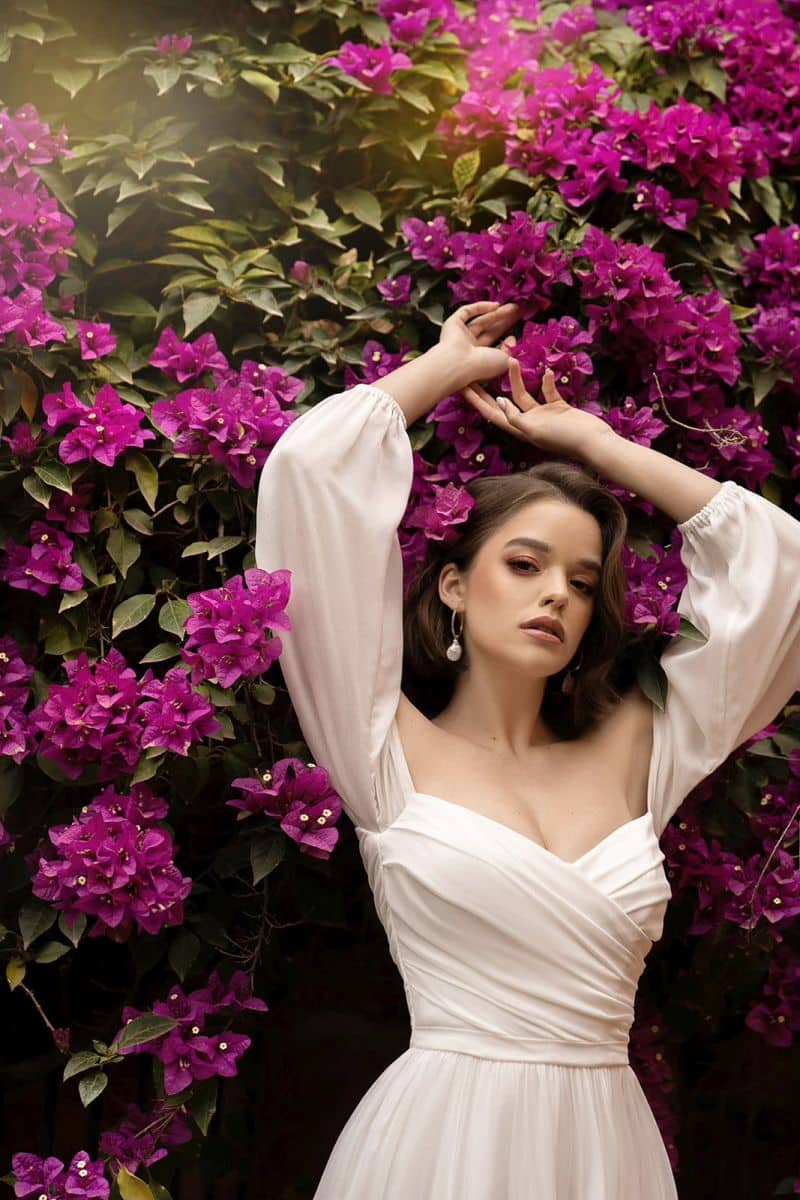 Eine Frau, die das Dama Couture Pola Hochzeitskleid trägt, posiert mit erhobenen Armen vor einer üppigen Wand aus leuchtend lila Bougainvillea-Blüten.