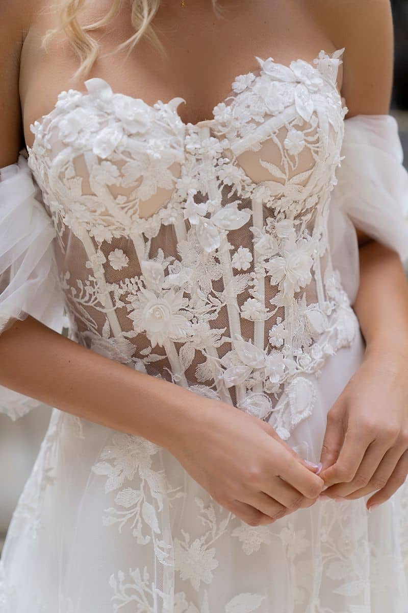 Nahaufnahme einer Frau, die das Dama Couture Sarah Brautkleid trägt, ein schulterfreies weißes Kleid mit Blumenapplikationen, durchsichtigem Stoff, zarten Tüllärmeln und aufwendigen Stickereien - ideal für Liebhaber eleganter Brautmoden.