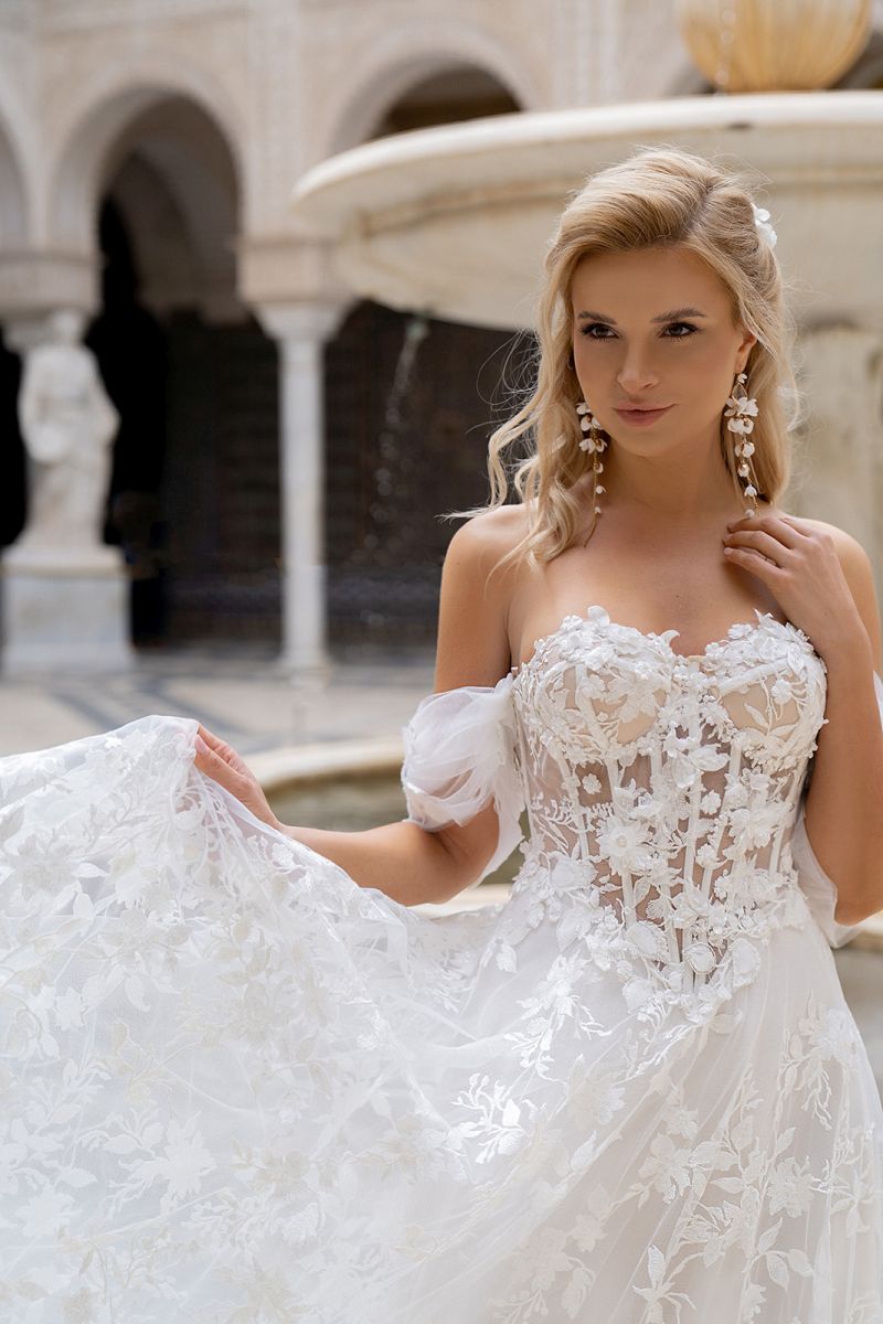 Eine Frau im Dama Couture Sarah Brautkleid steht vor einem Steinbrunnen und präsentiert anmutig ihr Kleid. Mit langen blonden Haaren und Statement-Ohrringen strahlt sie elegante Brautinspiration für moderne Brautmoden aus.