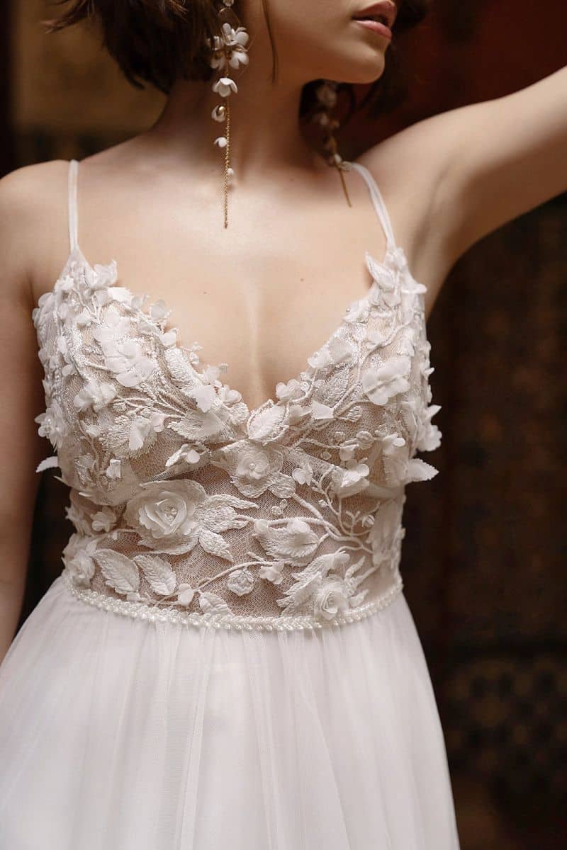 Eine Frau trägt das Dama Couture Tabita Hochzeitskleid, ein ärmelloses weißes Brautkleid mit dünnen Trägern und einem durchsichtigen Mieder aus floraler Spitze mit 3D-Blumenapplikationen; sie hat schulterlanges Haar und lange Ohrringe.
