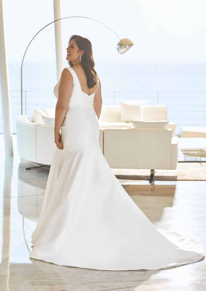 Eine Frau im Adoray Brautkleid von Pronovias - einem weißen, ärmellosen Kleid mit langer Schleppe - steht in einem Innenraum und blickt in die Ferne. Moderne weiße Möbel und große Fenster mit Blick aufs Meer schaffen eine elegante Hochzeitsszene.