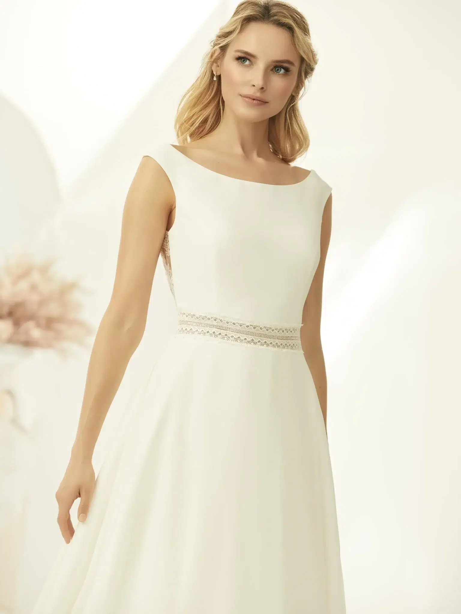 Eine Frau im Bianco Evento Parma Brautkleid, einem elfenbeinfarbenen ärmellosen Kleid mit Bateau-Ausschnitt und Spitzendetail an der Taille, steht vor einem sanft beleuchteten neutralen Hintergrund und verkörpert zeitlose Eleganz.