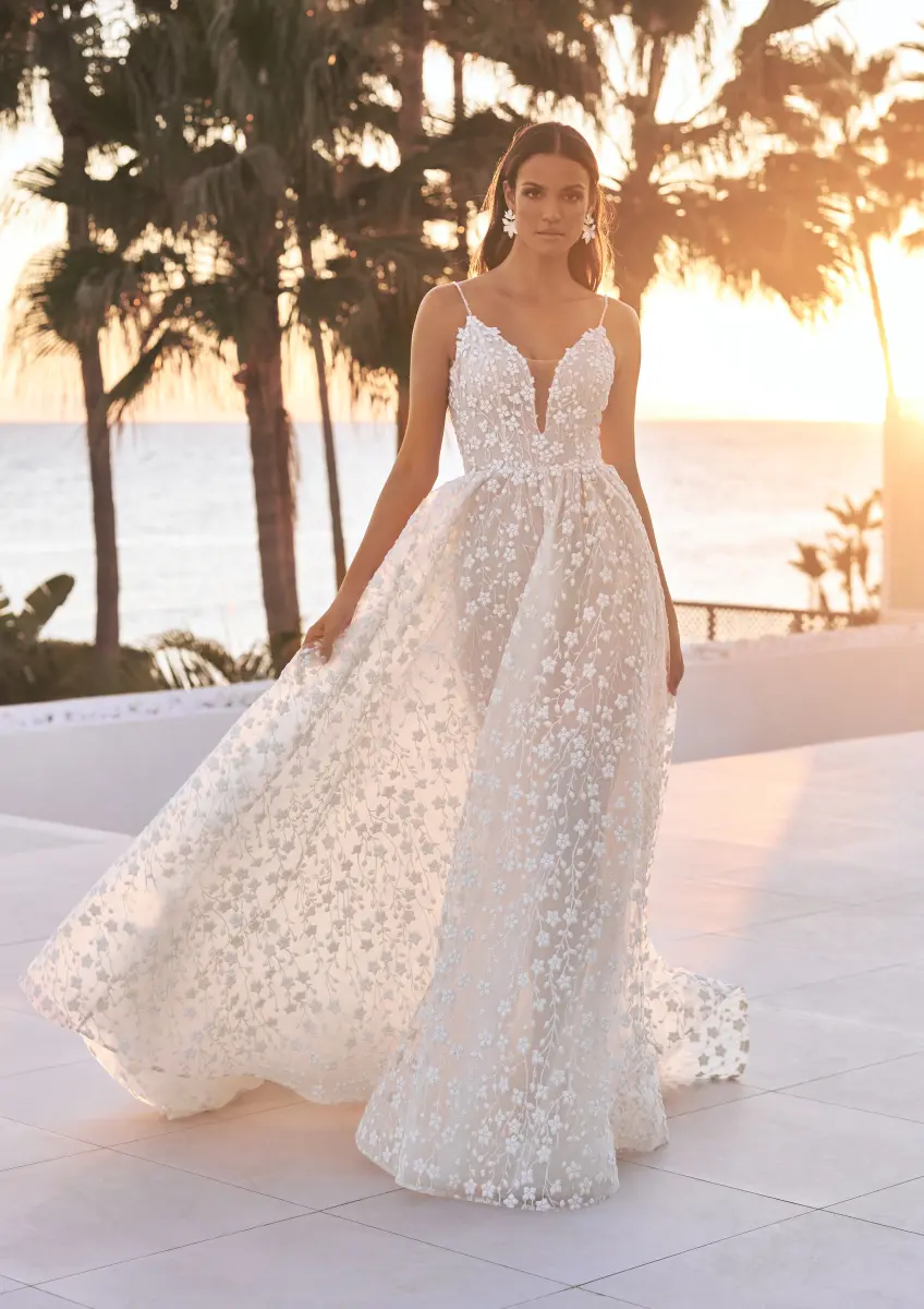 Eine Frau steht bei Sonnenuntergang im Freien und trägt das Yuki Brautkleid von Pronovias, ein ärmelloses Hochzeitskleid aus floraler Spitze mit tiefem Ausschnitt und fließendem, halbdurchsichtigem Rock, mit Palmen und dem Meer im Hintergrund.