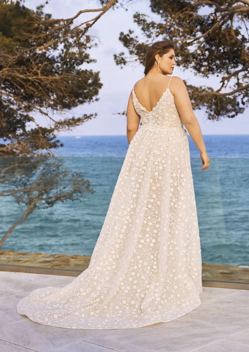 Eine Frau im ärmellosen, weißen Pronovias Yuki Brautkleid aus floraler Spitze mit langer Schleppe steht auf einer Marmorterrasse und blickt über ihre Schulter auf das blaue Meer und die Bäume. Entdecken Sie zeitlose Eleganz für Ihren besonderen Tag.
