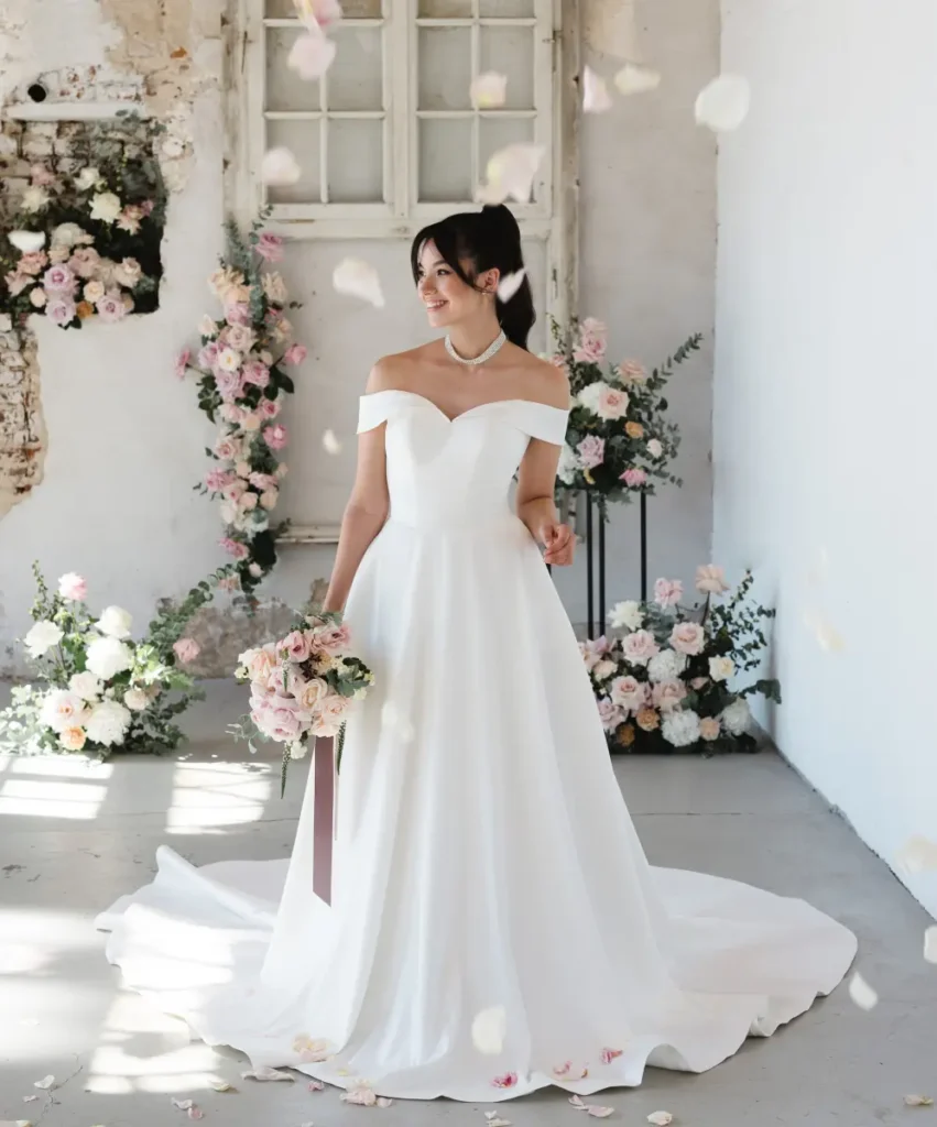 hochzeitsrausch Brautmoden - Elegantes Brautkleid mit schulterfreiem Design und Blumenstrauß in romantischem Setting