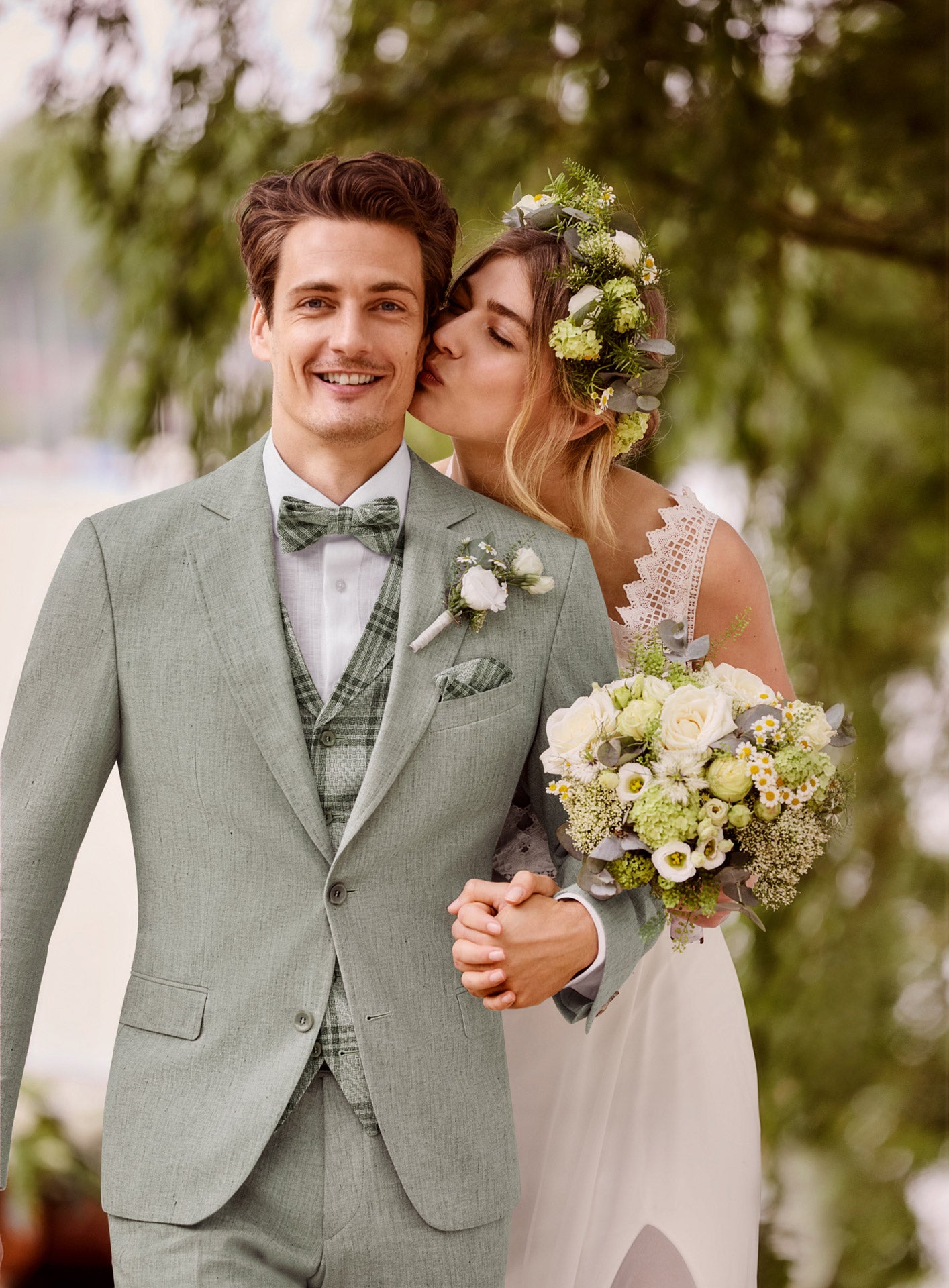 Eine Braut in einem weißen Kleid und einer Blumenkrone küsst die Wange ihres lächelnden Bräutigams. Er trägt das Atelier Torino Vintage Wedding Sakko 811773/40 mit einer Fliege, und sie halten sich im Freien inmitten von üppigem Grün die Hand, während sie einen Strauß weißer und grüner Blumen trägt.