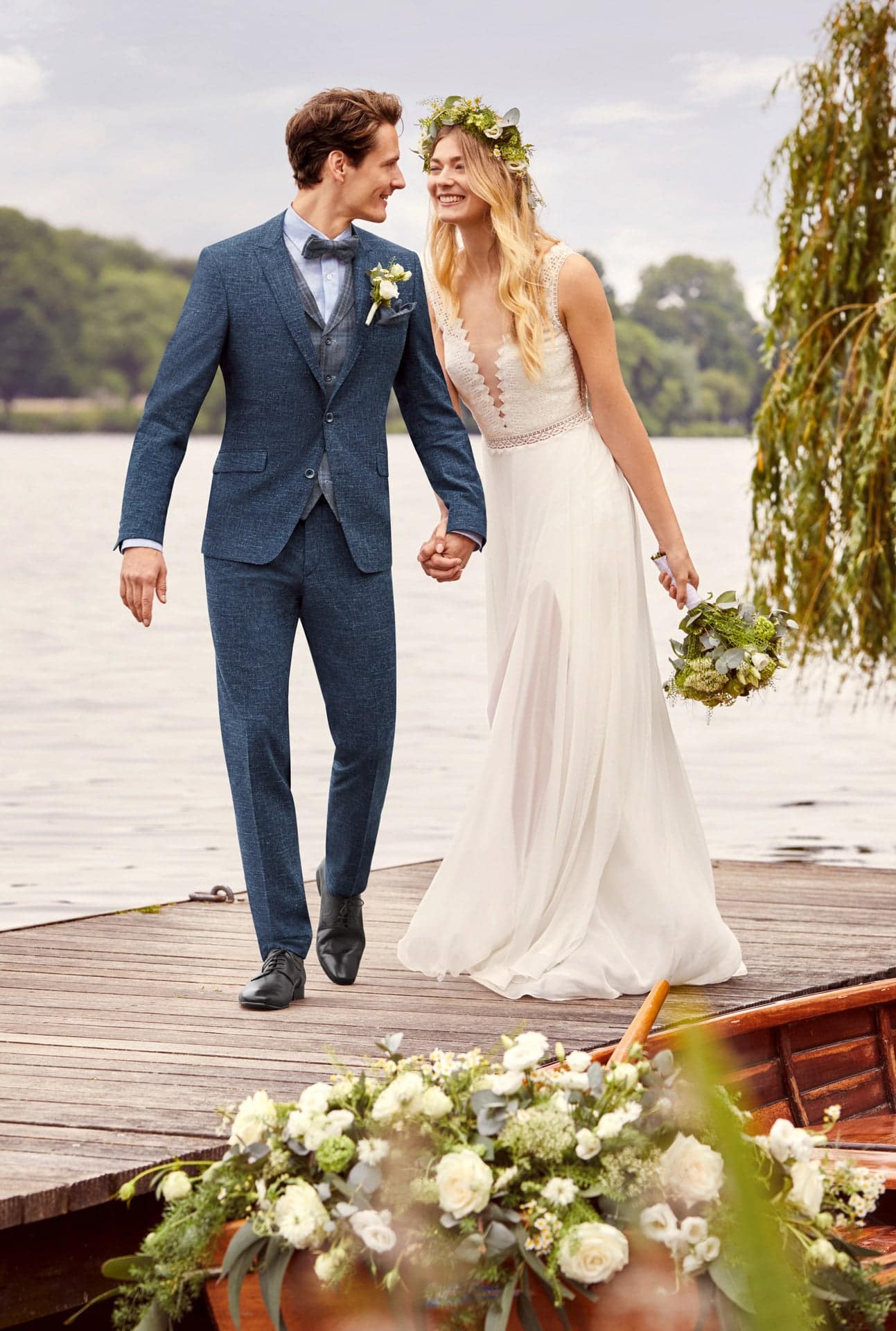 Der Bräutigam trägt das Atelier Torino Vintage Wedding Sakko 811775/30 und steht Hand in Hand mit seiner Braut in einem wunderschönen Hochzeitskleid und einer Blumenkrone an einem Seeufer. Weiße Blumen verleihen dem besonderen Moment Eleganz.