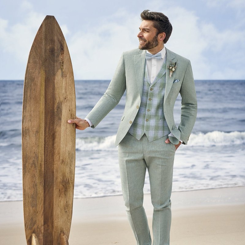 Ein Mann, der die Atelier Torino Vintage Wedding Weste 821986/40 trägt, steht lächelnd an einem Strand mit einem hölzernen Surfbrett - eine ideale Inspiration für einzigartige Brautmoden oder unkonventionelle Hochzeitskleid-Fotoshootings.