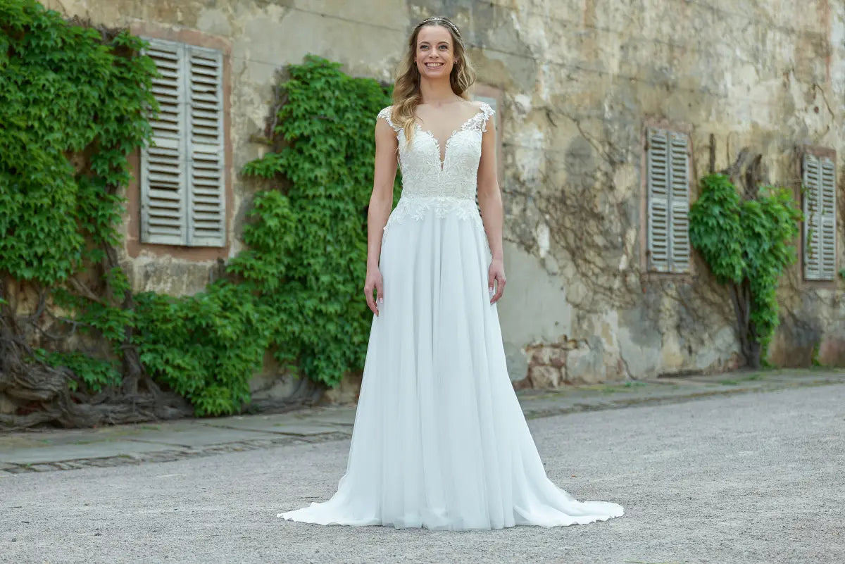 Eine Frau im Bridonista V24-22 Brautkleid steht lächelnd auf einem Kiesweg, mit efeubewachsenen, rustikalen Steinmauern und verschlossenen Fenstern im Hintergrund.