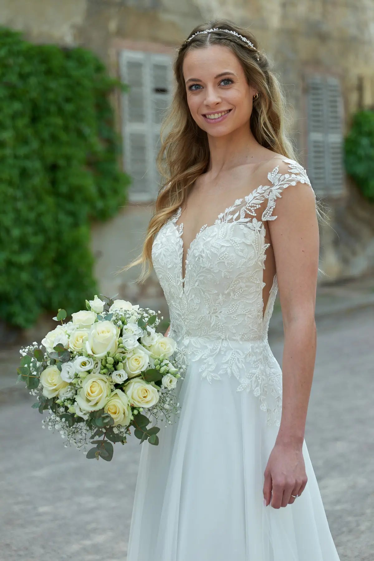 Eine lächelnde Braut trägt das Bridonista V24-22 Brautkleid und hält einen Strauß weißer und cremefarbener Rosen. Sie steht im Freien an einer Steinmauer mit grünem Efeu und verschlossenen Fenstern im Hintergrund.
