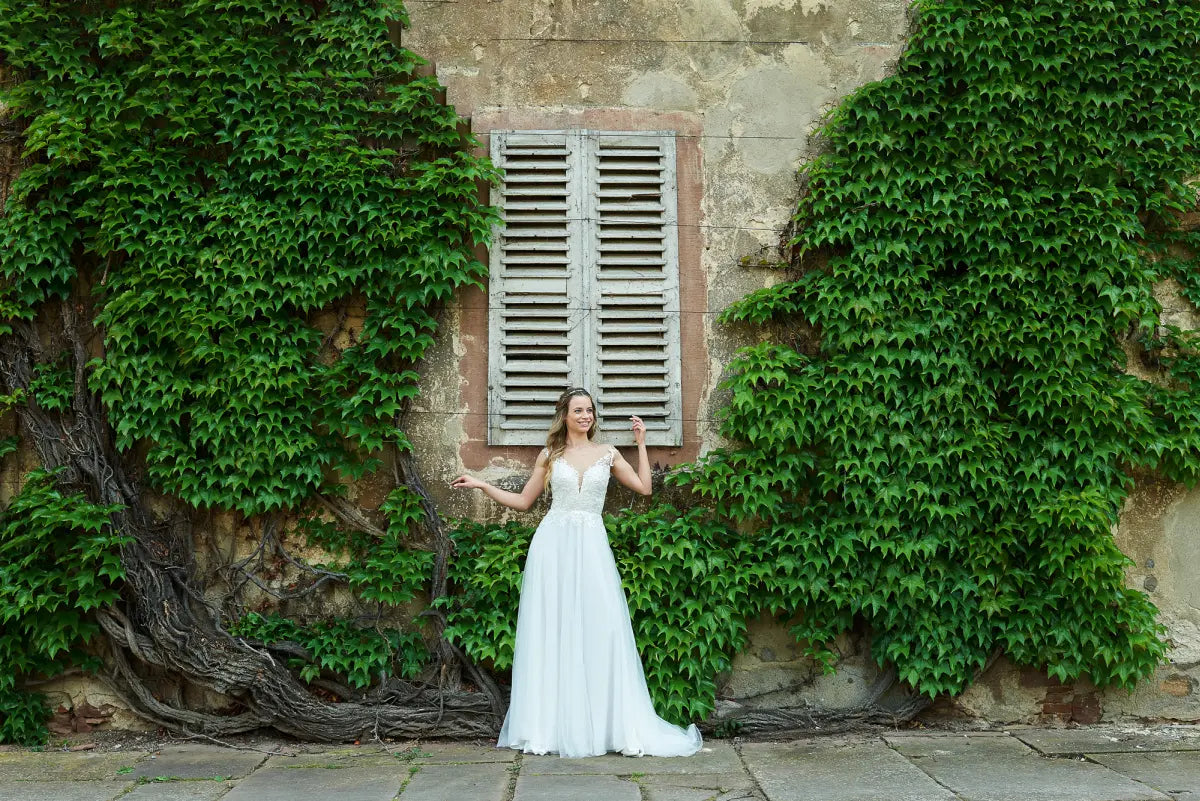 Eine Frau trägt das elegante Bridonista V24-22 Brautkleid und posiert mit leicht erhobenen Armen vor einer alten, efeubewachsenen Steinmauer neben einem geschlossenen, verwitterten Holzfenster.