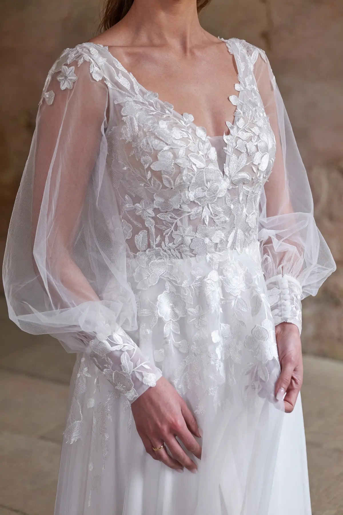 Eine Frau trägt das Bridonista V24-24 Brautkleid, ein weißes Hochzeitskleid mit durchsichtigen langen Puffärmeln und detaillierten floralen Spitzenapplikationen an Mieder und Ärmeln, vor einem neutralen Hintergrund.