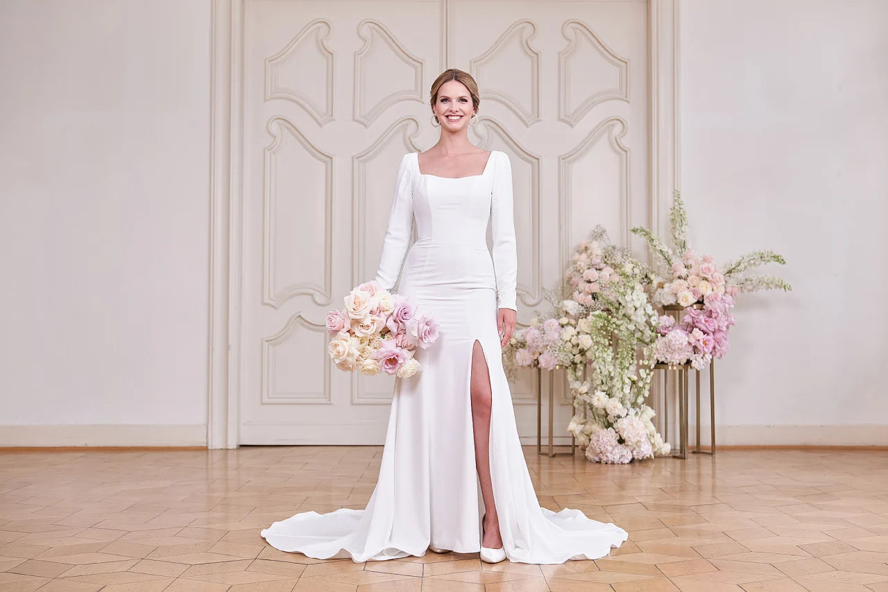 Eine Braut im Bridonista S24-05 Brautkleid mit langen Ärmeln und oberschenkelhohem Schlitz steht mit pastellfarbenen Blumen in der Hand vor verschnörkelten Flügeltüren und floralem Dekor - eine exquisite Wahl für moderne Brautmoden-Inspiration.