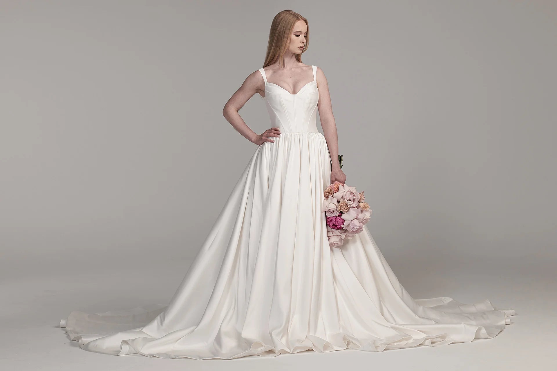 Eine Frau trägt das Bridonista BG24-24 Brautkleid, ein weißes, ärmelloses, bodenlanges Brautkleid, mit rosa und beigen Blumen vor einem schlichten hellgrauen Hintergrund.
