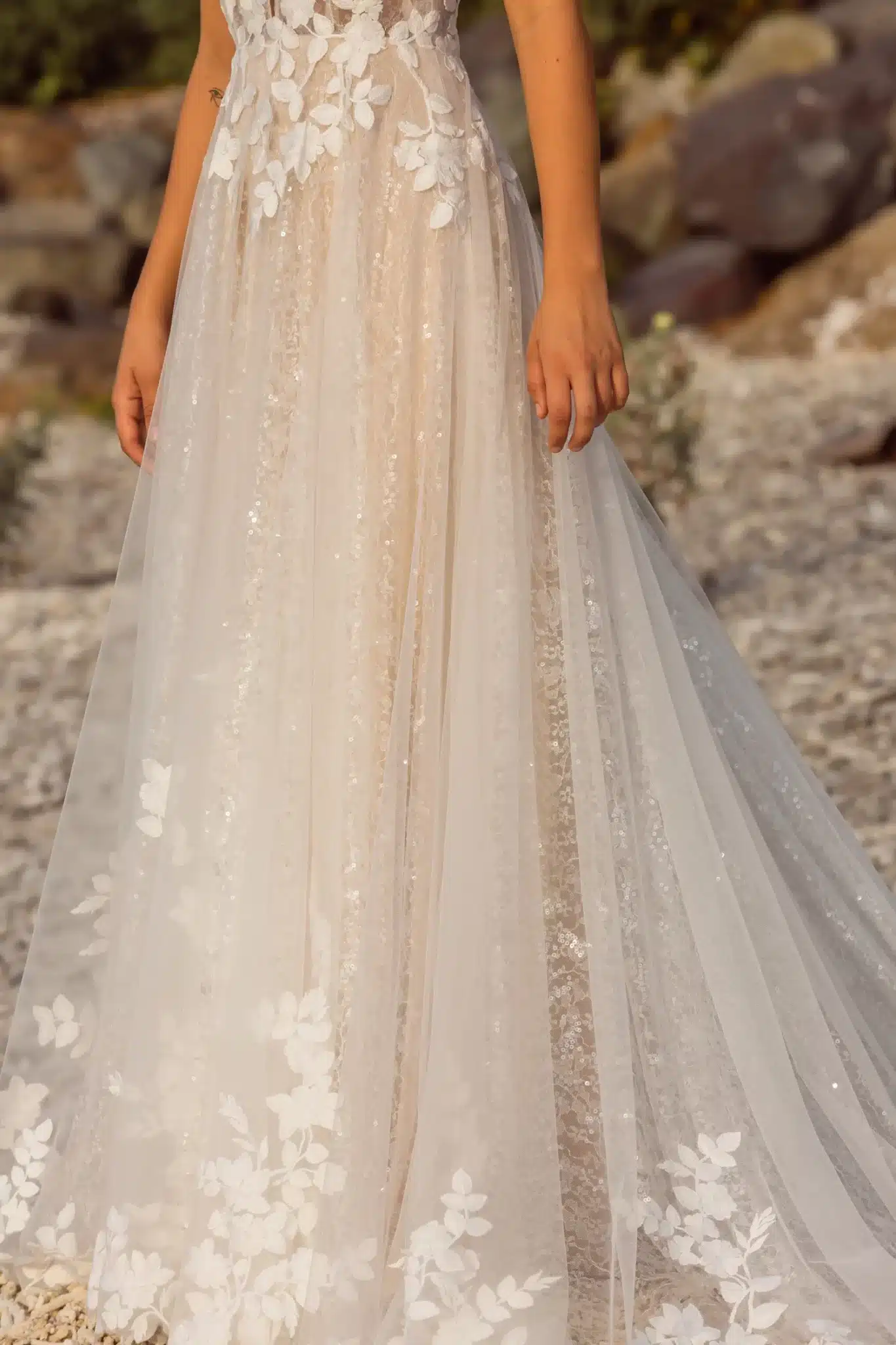 Eine Frau, die das Madi Lane Jenica Hochzeitskleid mit durchsichtigem Stoff, weißen Blumenapplikationen und Paillettenakzenten trägt, steht im Freien auf einer felsigen Fläche.