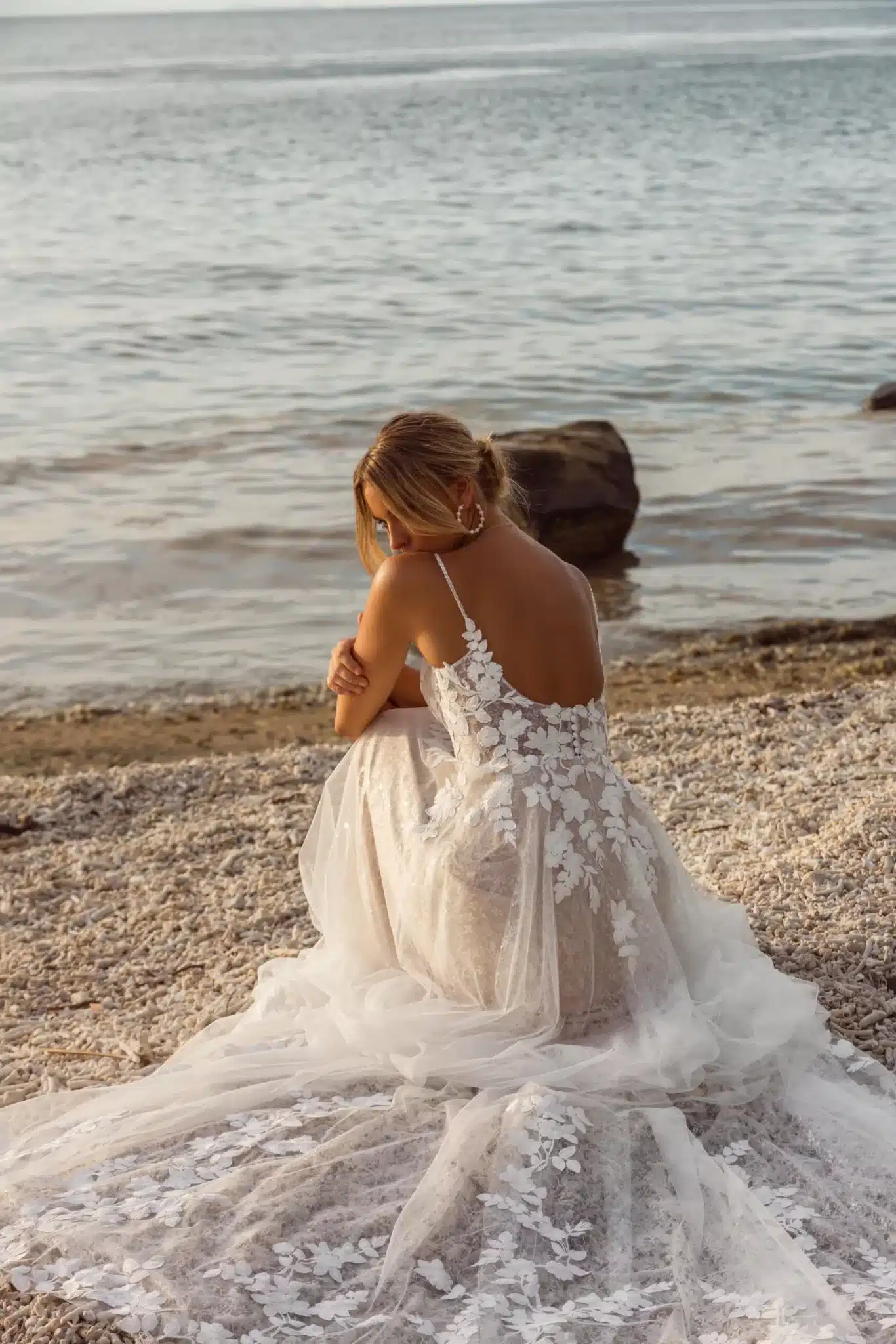 Eine Frau in einem weißen Madi Lane Jenica Hochzeitskleid aus geblümter Spitze sitzt auf einem Kiesstrand und blickt auf das ruhige Meer, den Kopf leicht gesenkt und das Haar zu einer lockeren Hochsteckfrisur frisiert.