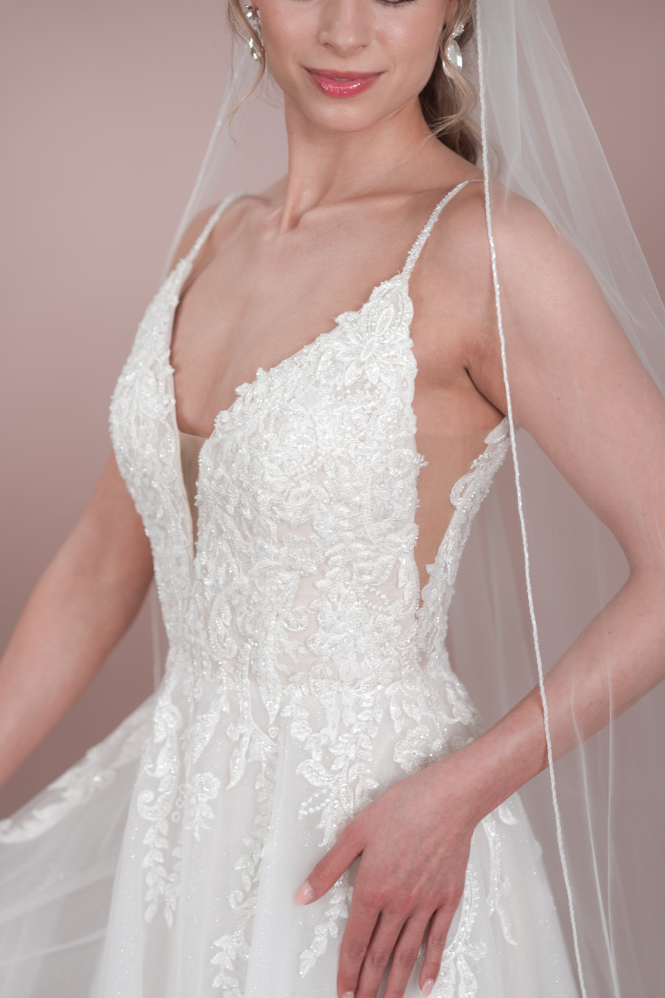 Eine Frau trägt das Bridonista C24-19 Hochzeitskleid, ein weißes Spitzenkleid mit dünnen Trägern, tiefem Ausschnitt und aufwändiger Stickerei, gestylt mit einem Schleier, hellrosa Lippenstift und Perlenohrringen.