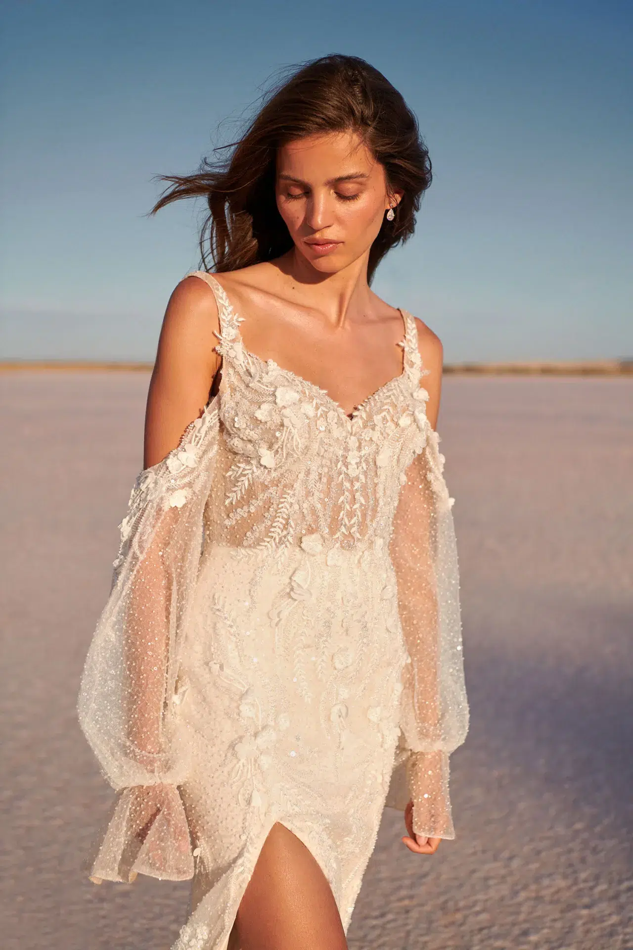 Eine Frau steht im Freien in dem Jeune Bridal Medina Hochzeitskleid, einem cremefarbenen Kleid mit durchsichtigen, wogenden Ärmeln und floralen Details. Die sandige Landschaft und der klare Himmel bieten eine atemberaubende Kulisse für dieses elegante Brautmodenstück.