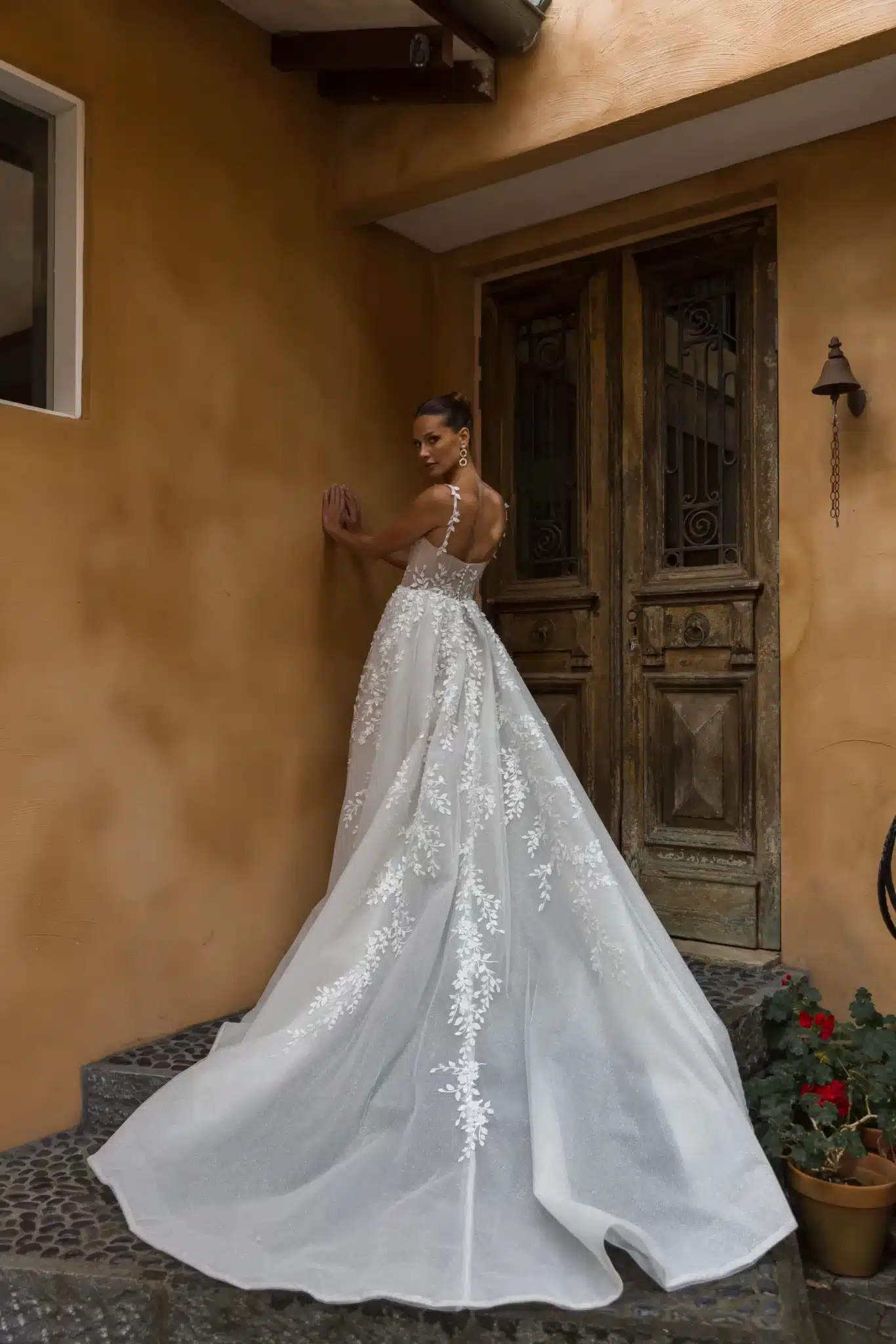 Eine Frau in dem Madi Lane Pia Brautkleid steht auf Steinstufen vor alten Holztüren. Die spitzenverzierte Schleppe und die durchsichtigen Träger des Kleides setzen den eleganten Rücken in Szene, während sie die Wand berührt und zeitlosen Brautcharme versprüht.
