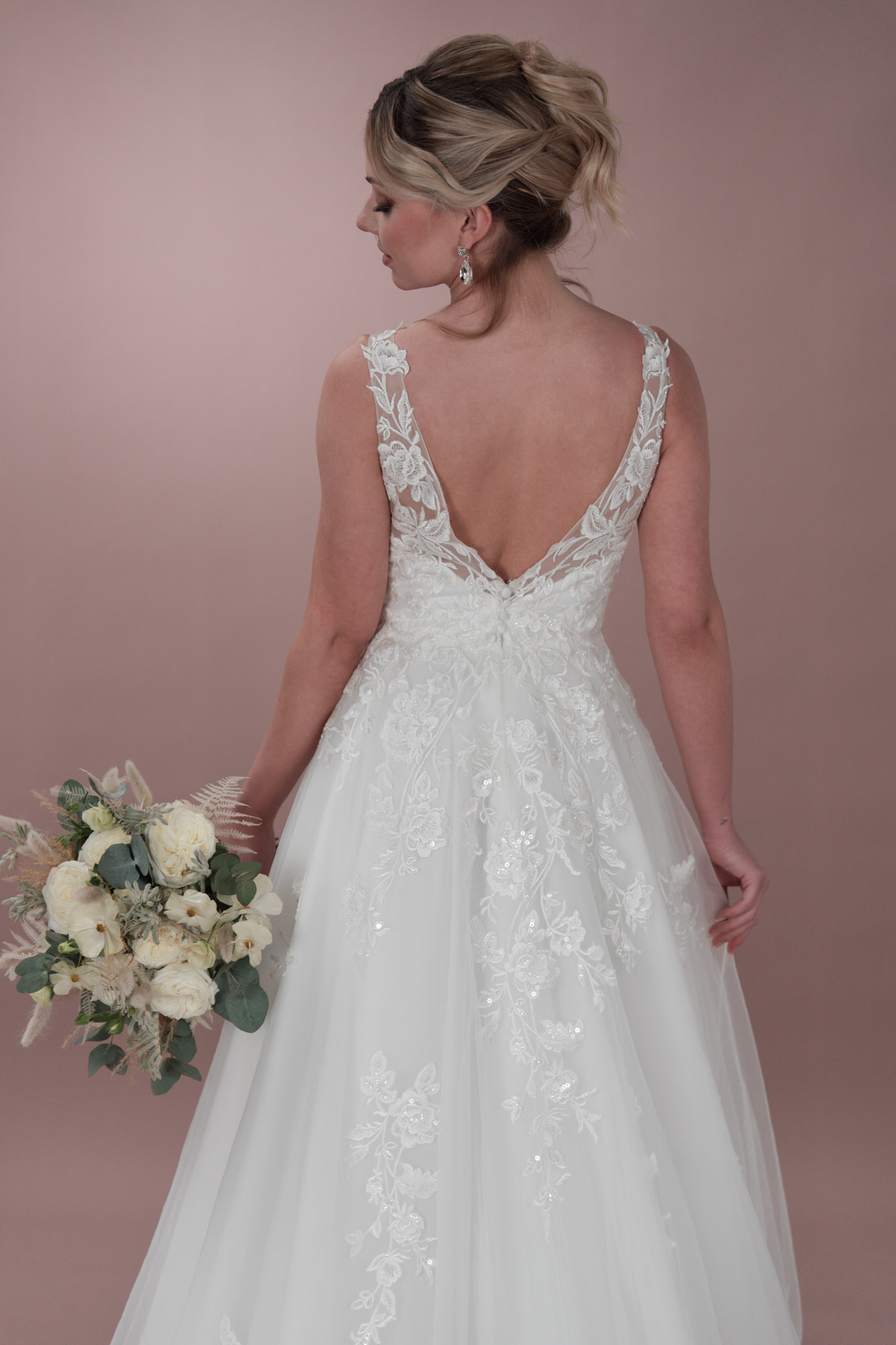 Eine Frau im Bridonista C24-23 Hochzeitskleid mit Spitzenstickerei steht mit dem Rücken zur Kamera und hält weiße Rosen in der Hand. Ihr blondes Haar ist vor einem zartrosa Hintergrund hochgesteckt - ein Beispiel für moderne Brautmoden-Eleganz.