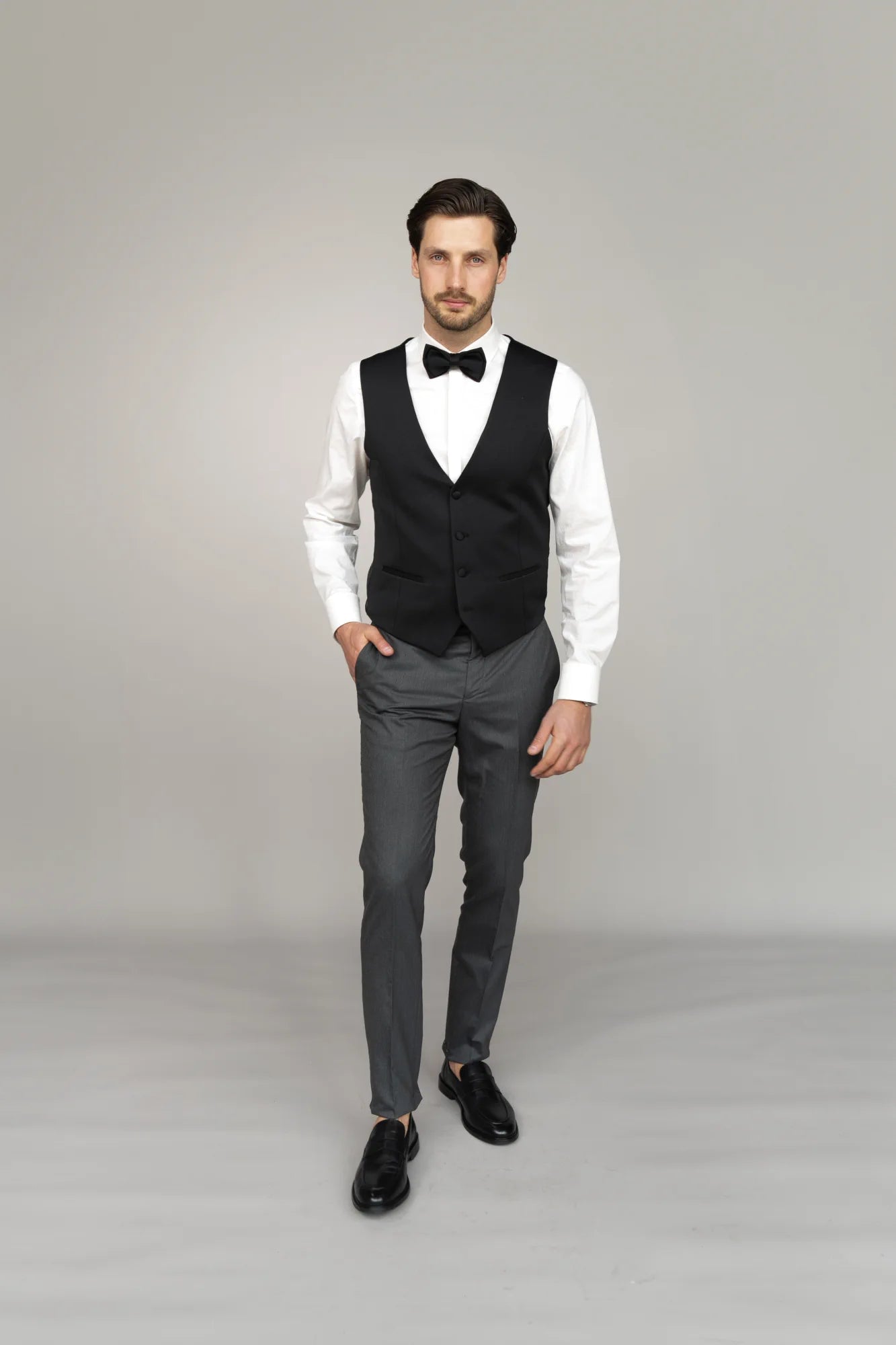 Ein Mann mit kurzen dunklen Haaren in formeller Kleidung, der Masculini 3397-08 trägt, steht vor einem grauen Hintergrund. Neben ihm bilden elegante Brautkleider und Hochzeitskleider den idealen Rahmen für ein stilvolles Hochzeitsensemble.