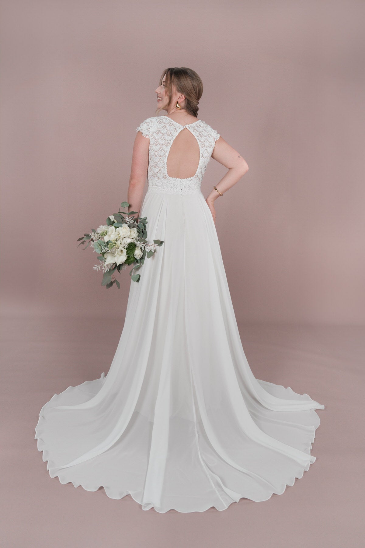 Eine Braut im Bridonista R2L25-C-A11 Hochzeitskleid mit Spitzenrücken und Schlüsselloch-Detail steht mit dem Rücken zur Wand und hält einen weißen Blumenstrauß. Das Kleid hat eine lange Schleppe, Kappenärmel und ist vor einem hellvioletten Hintergrund zu sehen.