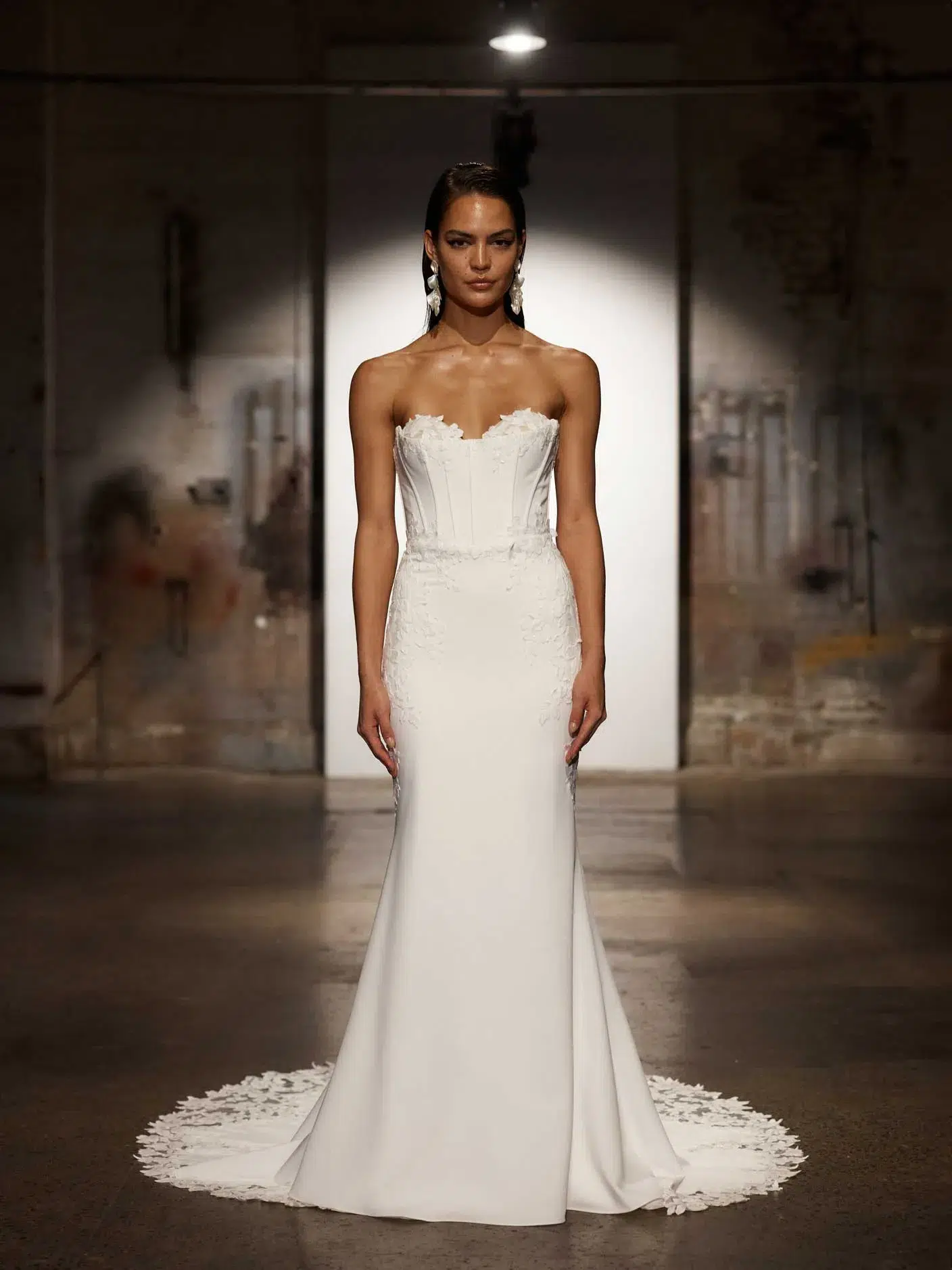 Eine Frau modelliert das Evie Young Hale, ein trägerloses, figurbetontes Hochzeitskleid mit Spitzendetails und einer leichten Schleppe, auf einem schwach beleuchteten Laufsteg im Industriestil. Sie vervollständigt ihren Look mit auffälligen Ohrringen.