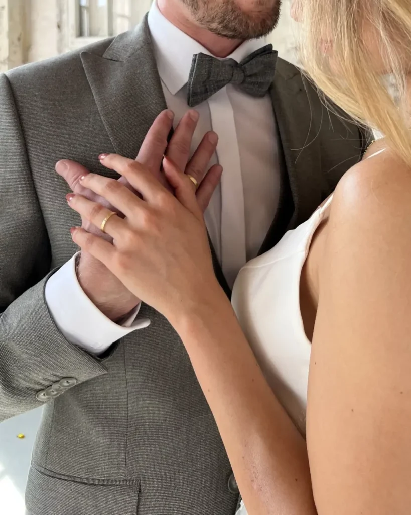 hochzeitsrausch Eheringe - Brautpaar in eleganter Umarmung, Braut im weißen Kleid, Bräutigam im grauen Anzug mit Fliege