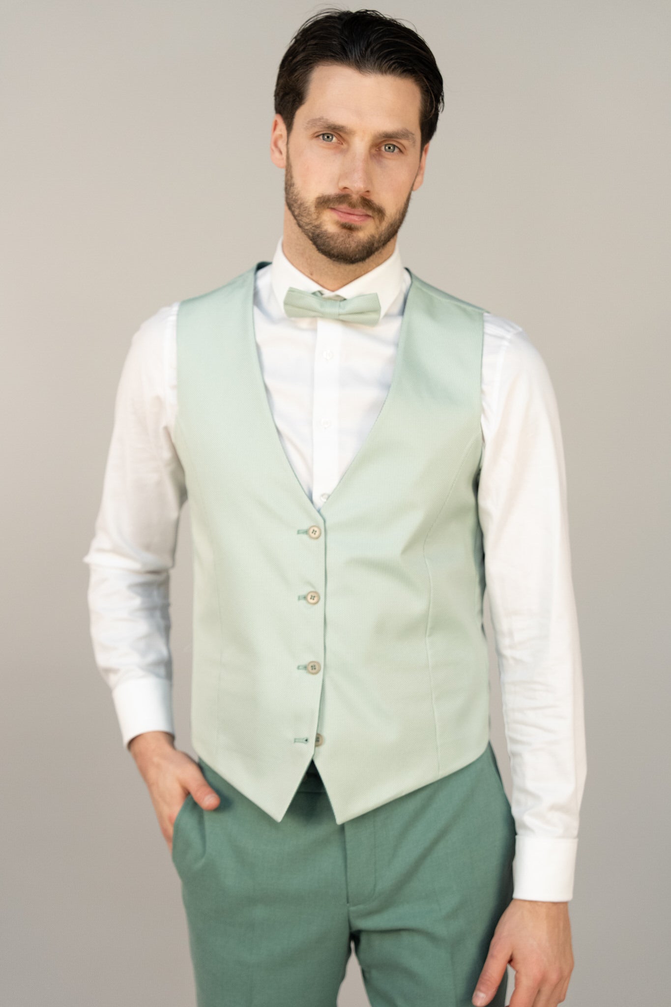 Ein Mann mit kurzen dunklen Haaren und Bart trägt selbstbewusst das Masculini 803-07A: eine hellgrüne Weste, eine passende Fliege und eine grüne Hose über einem weißen Hemd - ein elegantes Ensemble, das perfekt zu jedem Brautkleid passt.