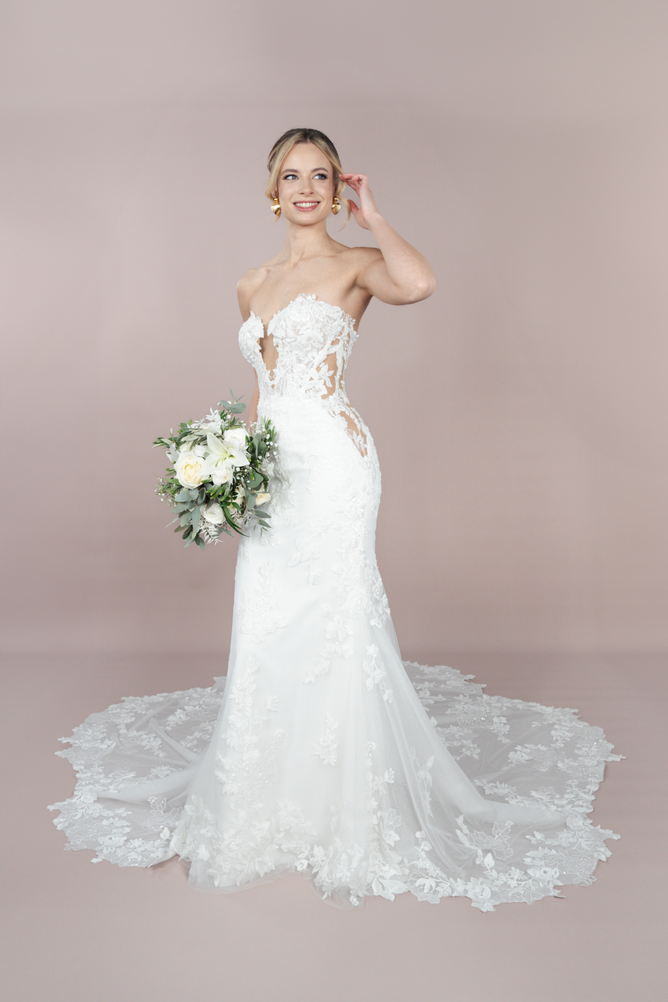 Eine Braut in dem trägerlosen Spitzenhochzeitskleid C25-F06 von Bridonista mit langer Schleppe hält einen Strauß weißer Blumen und Grünzeug vor einem zartrosa Hintergrund.