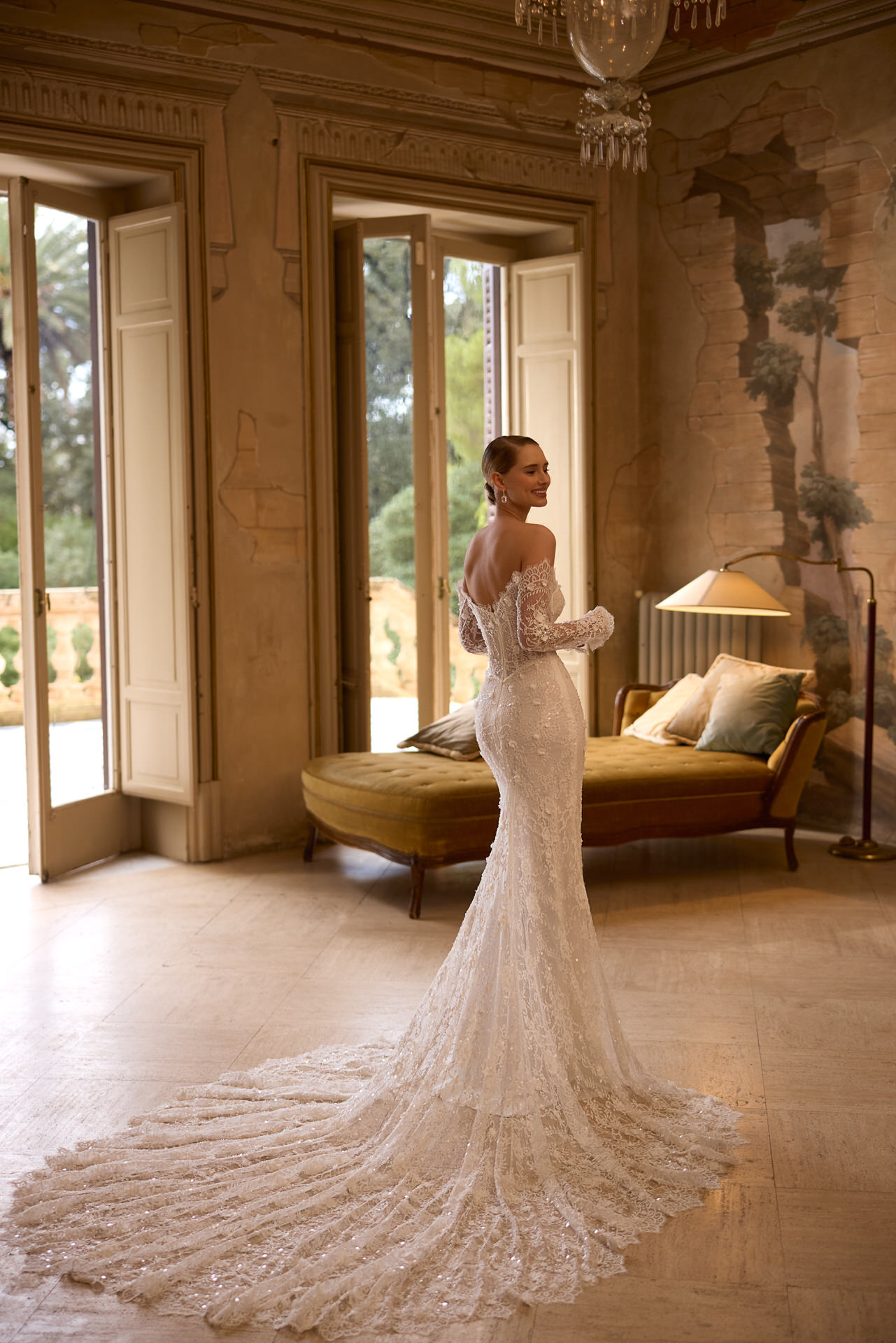 Eine Frau in einem eleganten, schulterfreien Elizabeth Passion 5817T Brautkleid mit langer Spitzenschleppe steht in einem raffinierten Raum mit hohen Fenstern, klassischem Dekor und einer alten Chaiselongue und blickt über ihre Schulter.