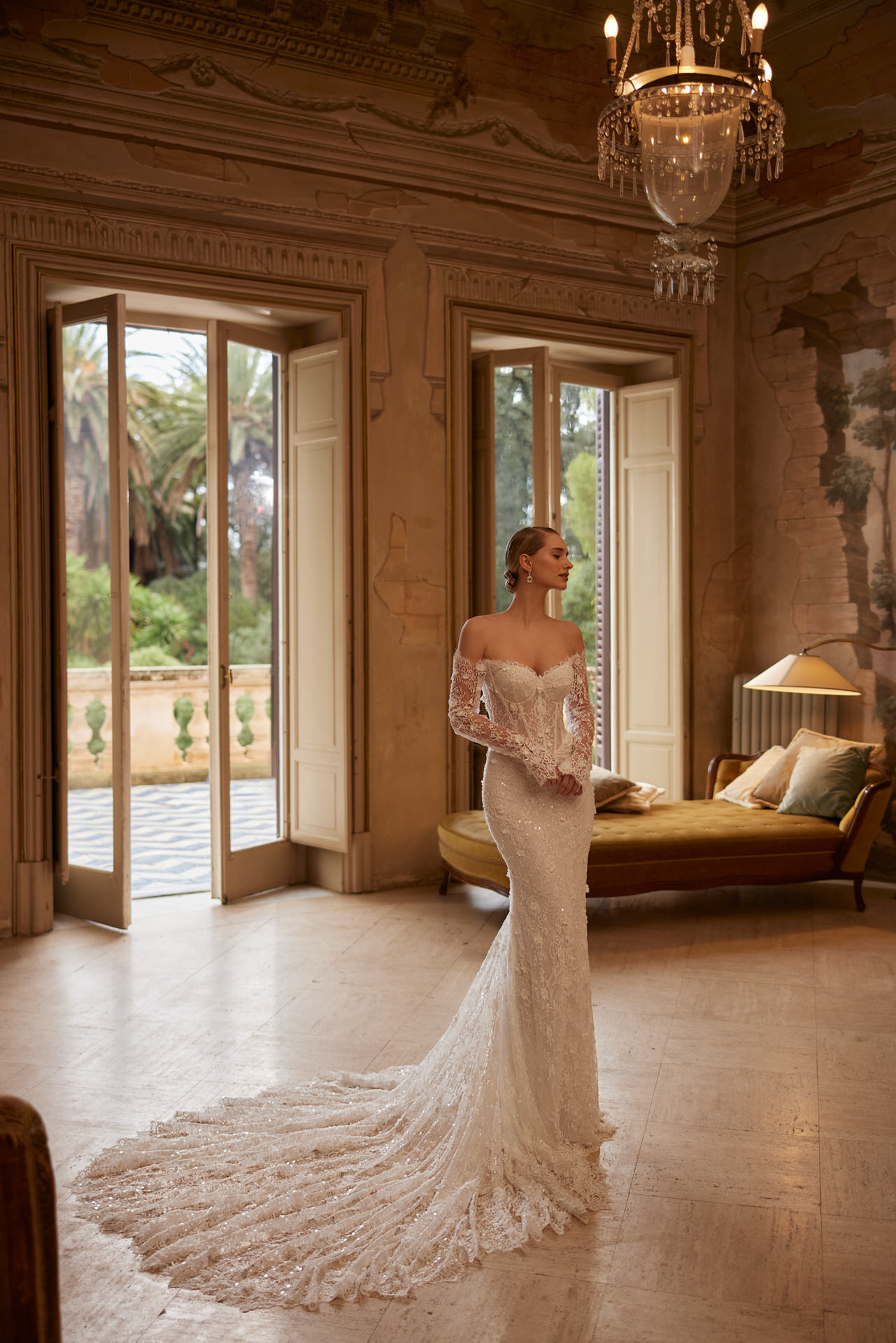 Eine Frau trägt das elegante Elizabeth Passion 5817T Brautkleid mit langer Schleppe, während sie in einem großen Vintage-Zimmer mit hohen Fenstern, einem Kronleuchter und Blick auf den Garten steht.