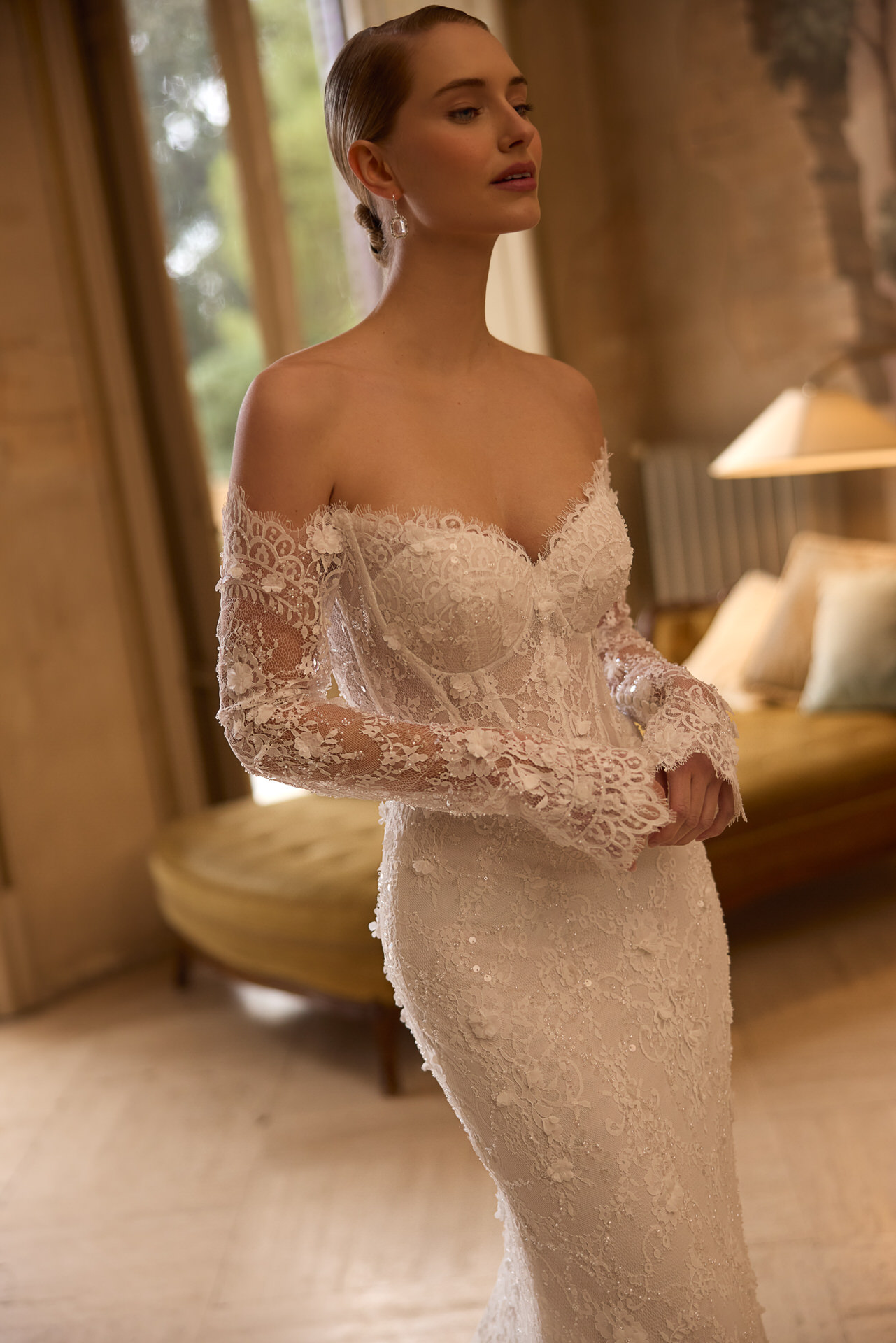 Eine Frau im Elizabeth Passion 5817T Brautkleid, einem schulterfreien, langärmeligen Spitzenhochzeitskleid, steht an einem von natürlichem Licht beleuchteten Fenster; im Hintergrund sind eine grüne Couch und eine Lampe zu sehen.