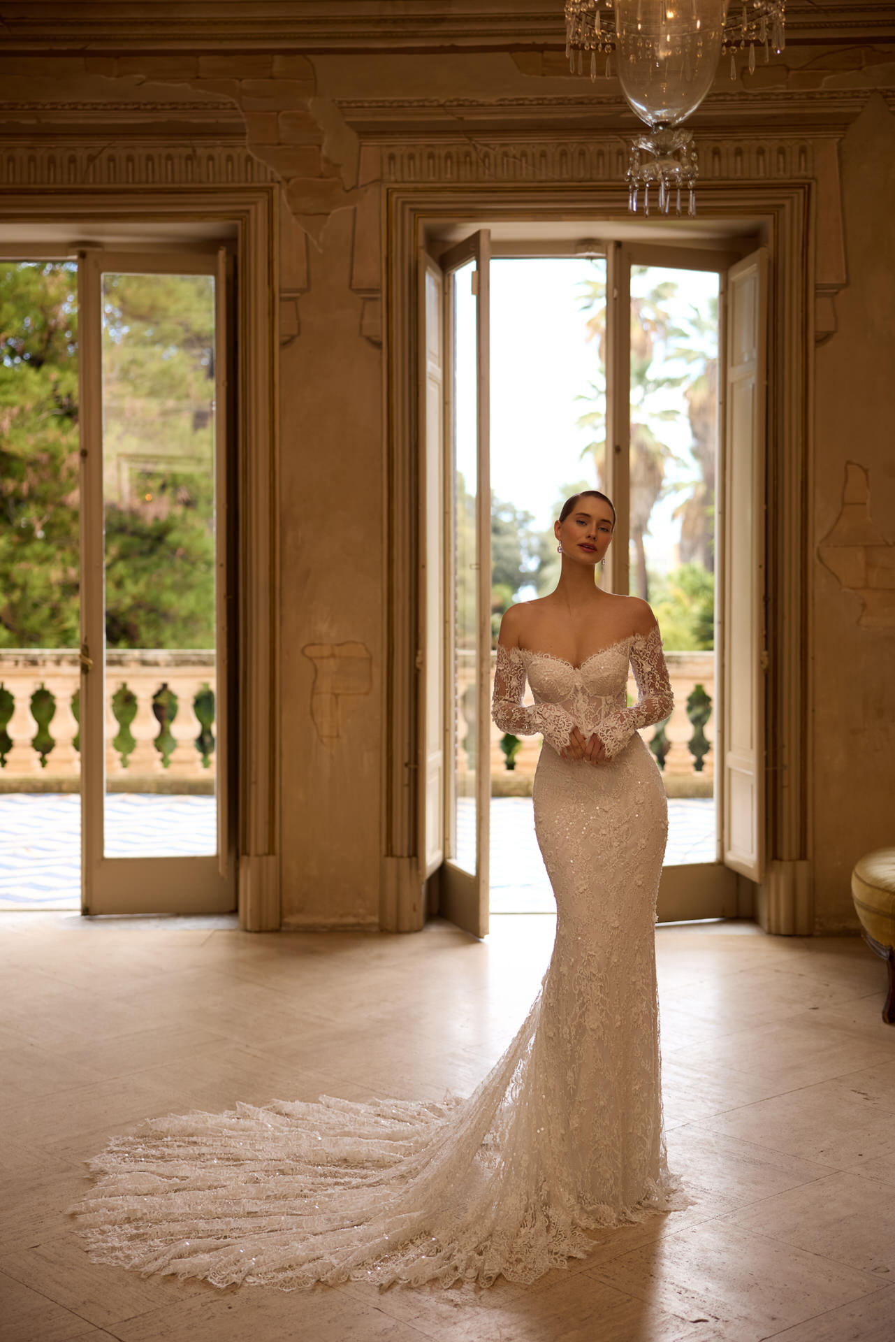 Eine Frau in einem eleganten Elizabeth Passion 5817T Brautkleid mit schulterfreiem Schnitt und langer Schleppe steht in einem sonnendurchfluteten Raum vor hohen offenen Türen und blickt auf einen Balkon inmitten von üppigem Grün.