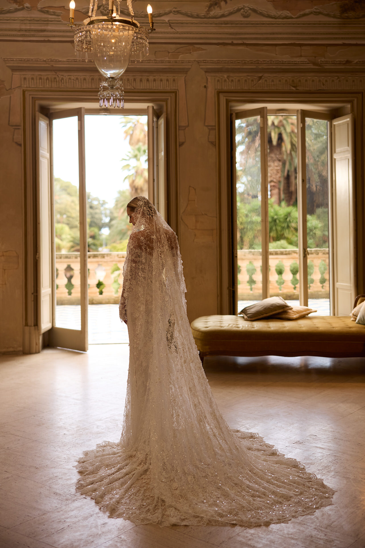 Eine Braut in einem Elizabeth Passion 5817T Brautkleid und Schleier steht in einem hellen, eleganten Raum mit hohen Decken und großen Fenstern und blickt auf das üppige Grün hinaus.