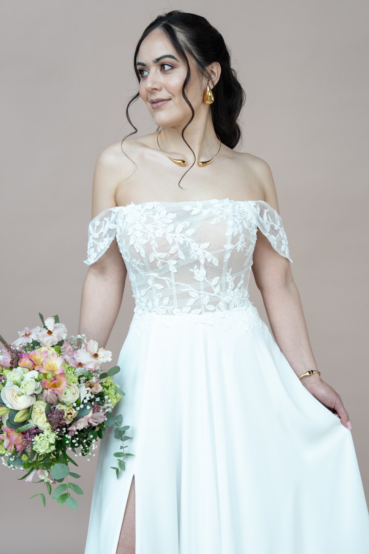 Eine Frau in dem schulterfreien, spitzenbesetzten Hochzeitskleid Bridonista V26-A01 hält einen Strauß mit rosa, weißen und gelben Blumen. Sie hat langes, dunkles, gewelltes Haar, trägt Goldschmuck und steht vor einem hellbeigen Hintergrund.