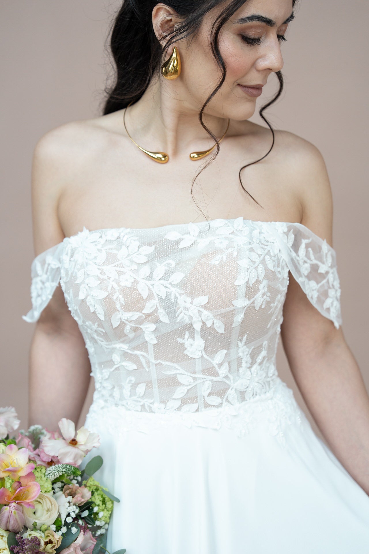 Eine Frau in dem schulterfreien, geblümten Spitzenkleid Bridonista V26-A01 hält einen Blumenstrauß und trägt Goldschmuck. Ihr langes dunkles Haar ist zu lockeren Wellen gestylt und sie blickt mit einem sanften Lächeln zu Boden.