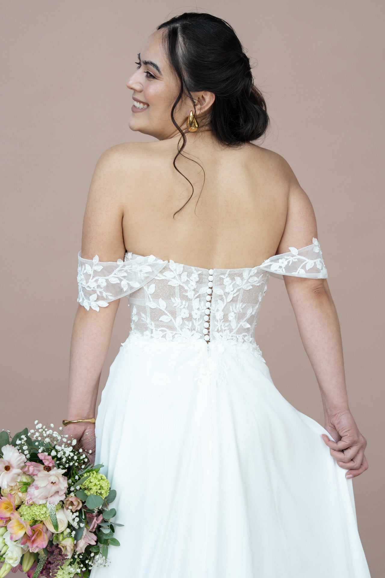 Eine lächelnde Braut blickt von sich weg und zeigt die Rückseite ihres schulterfreien Bridonista V26-A01 Brautkleids aus Spitze. Sie hält einen bunten Blumenstrauß in der Hand und blickt über ihre Schulter vor einem einfarbig beigen Hintergrund.