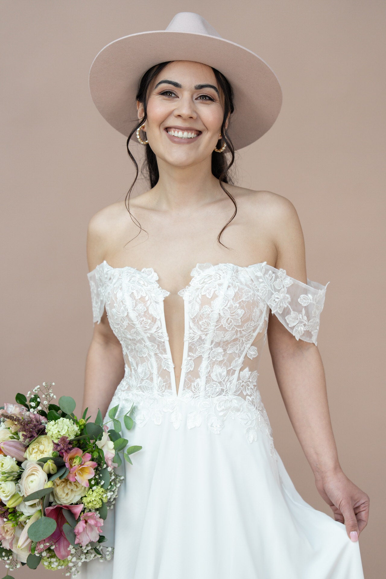Eine lächelnde Braut im schulterfreien Brautkleid Bridonista V26-A02 und einem breitkrempigen Hut hält einen bunten Blumenstrauß vor einem schlichten beigen Hintergrund.