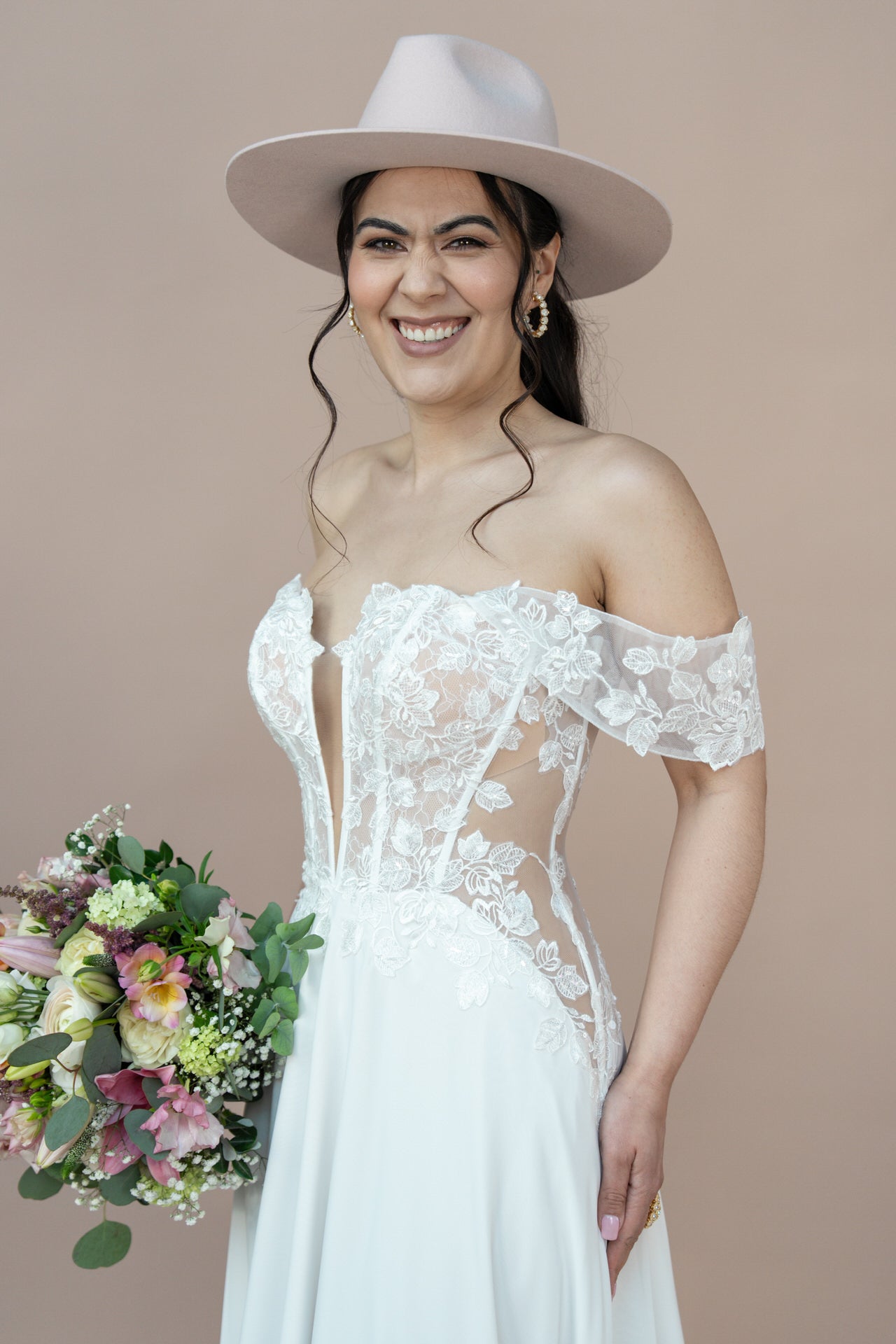 Eine lächelnde Frau im schulterfreien Brautkleid Bridonista V26-A02 und einem hellrosa Hut hält einen bunten Blumenstrauß vor einem schlichten beigen Hintergrund.