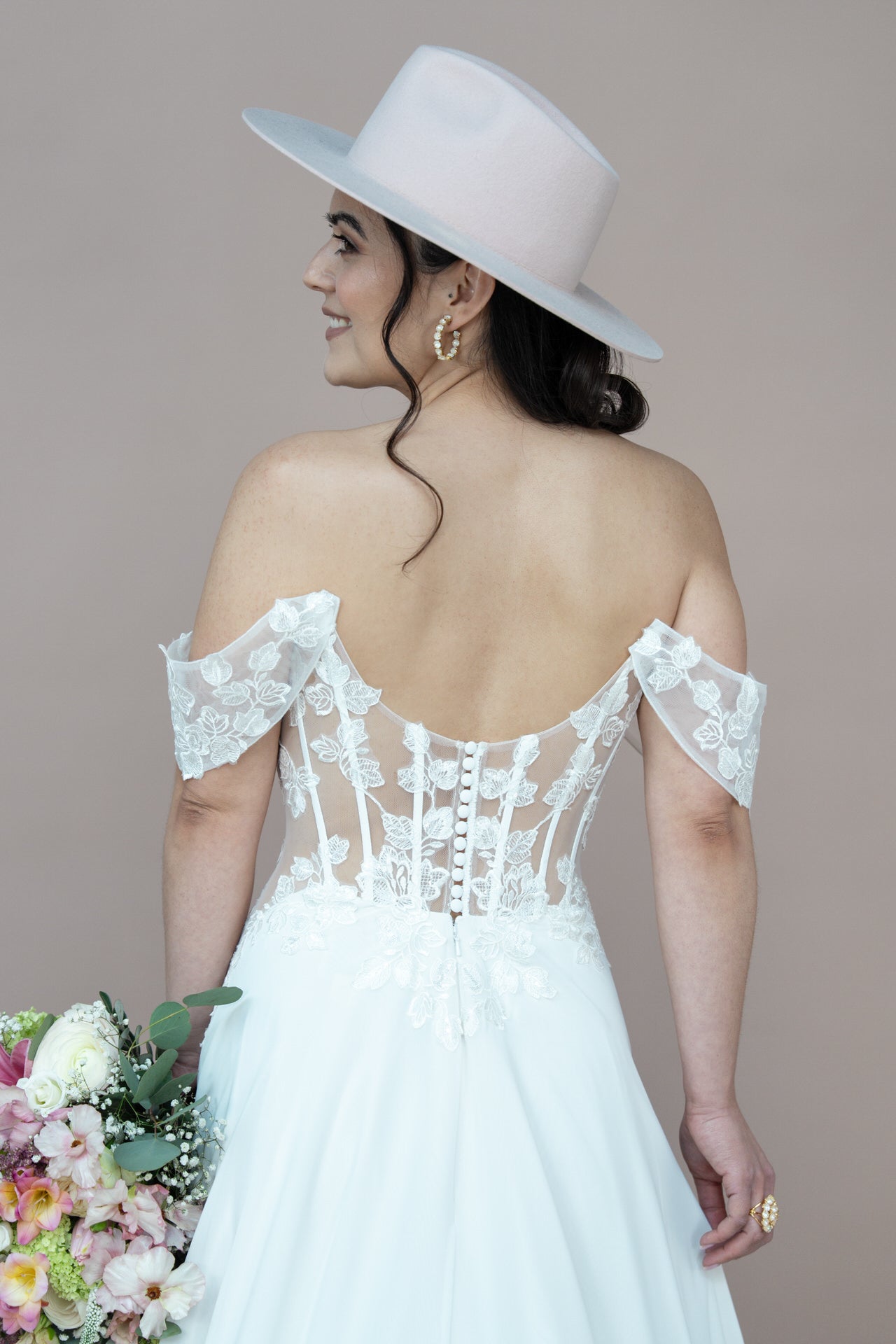 Eine Braut, die das schulterfreie Spitzenkleid Bridonista V26-A02 und einen breitkrempigen hellen Hut trägt, blickt nach vorne und hält einen pastellfarbenen Blumenstrauß. Ihr dunkles Haar ist in lockere Wellen gelegt und sie trägt goldene Ohrringe.