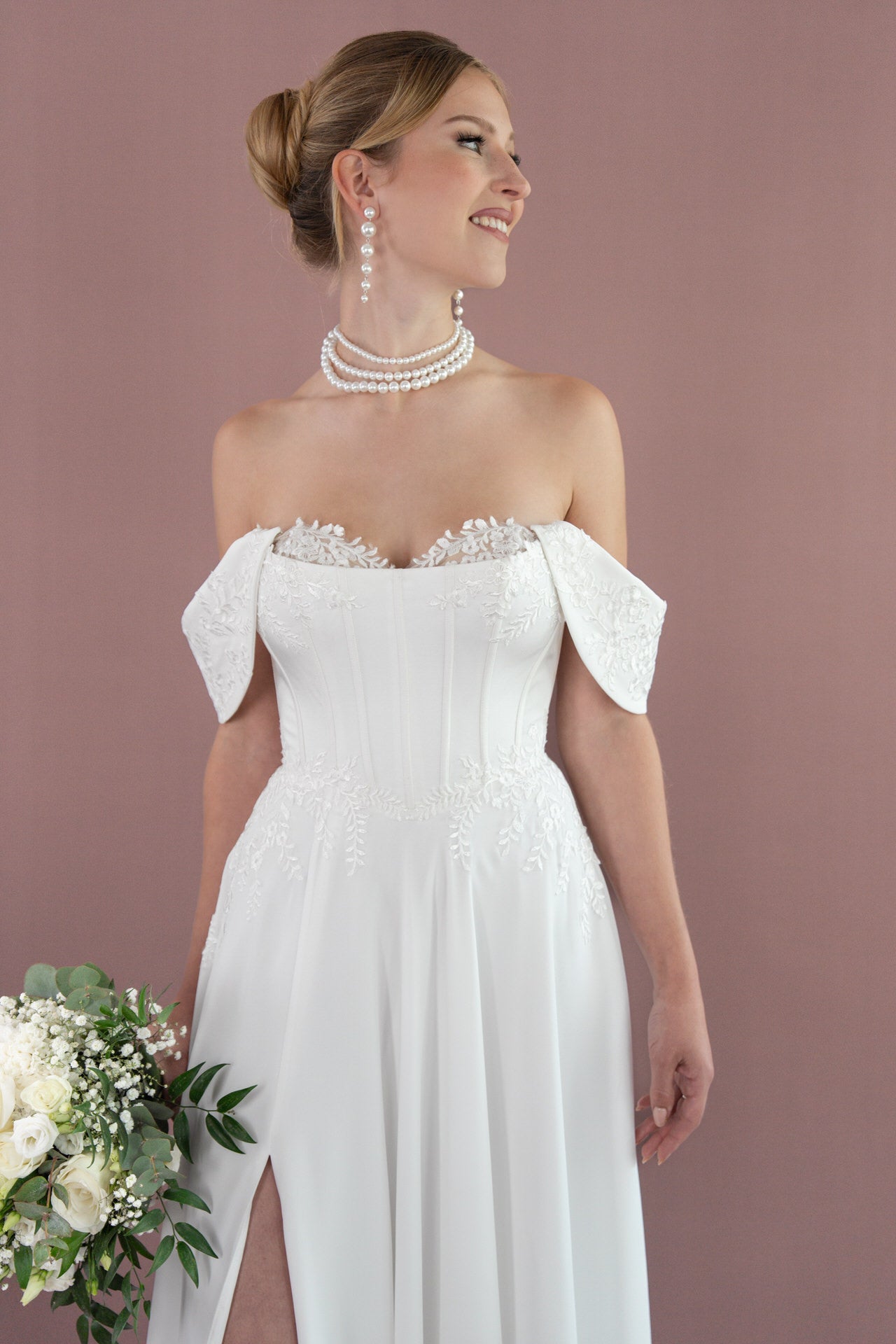 Eine fröhliche Braut trägt das schulterfreie weiße Brautkleid Bridonista V26-A03 mit floralen Details und hält einen weiß-grünen Strauß. Perlenschmuck und ein mauvefarbener Hintergrund unterstreichen ihre zeitlose Brautmoden-Eleganz.