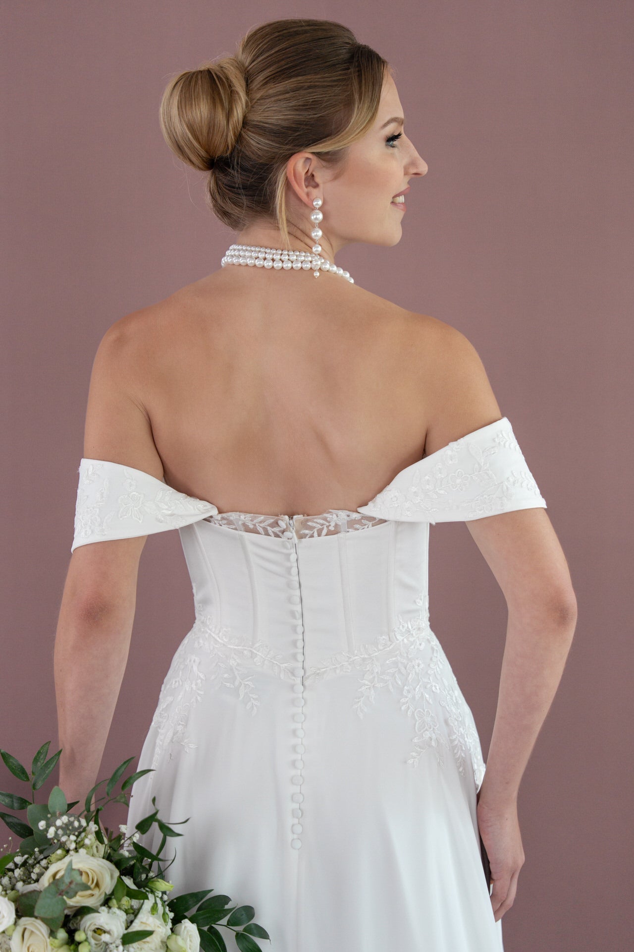 Eine Frau im schulterfreien Hochzeitskleid Bridonista V26-A03 mit Spitzendetails steht mit dem Rücken zugewandt und hält einen Strauß weißer Rosen und Grünzeug in der Hand. Ihr Haar ist zu einem eleganten Dutt gebunden und mit Perlenohrringen und einer Perlenkette geschmückt.